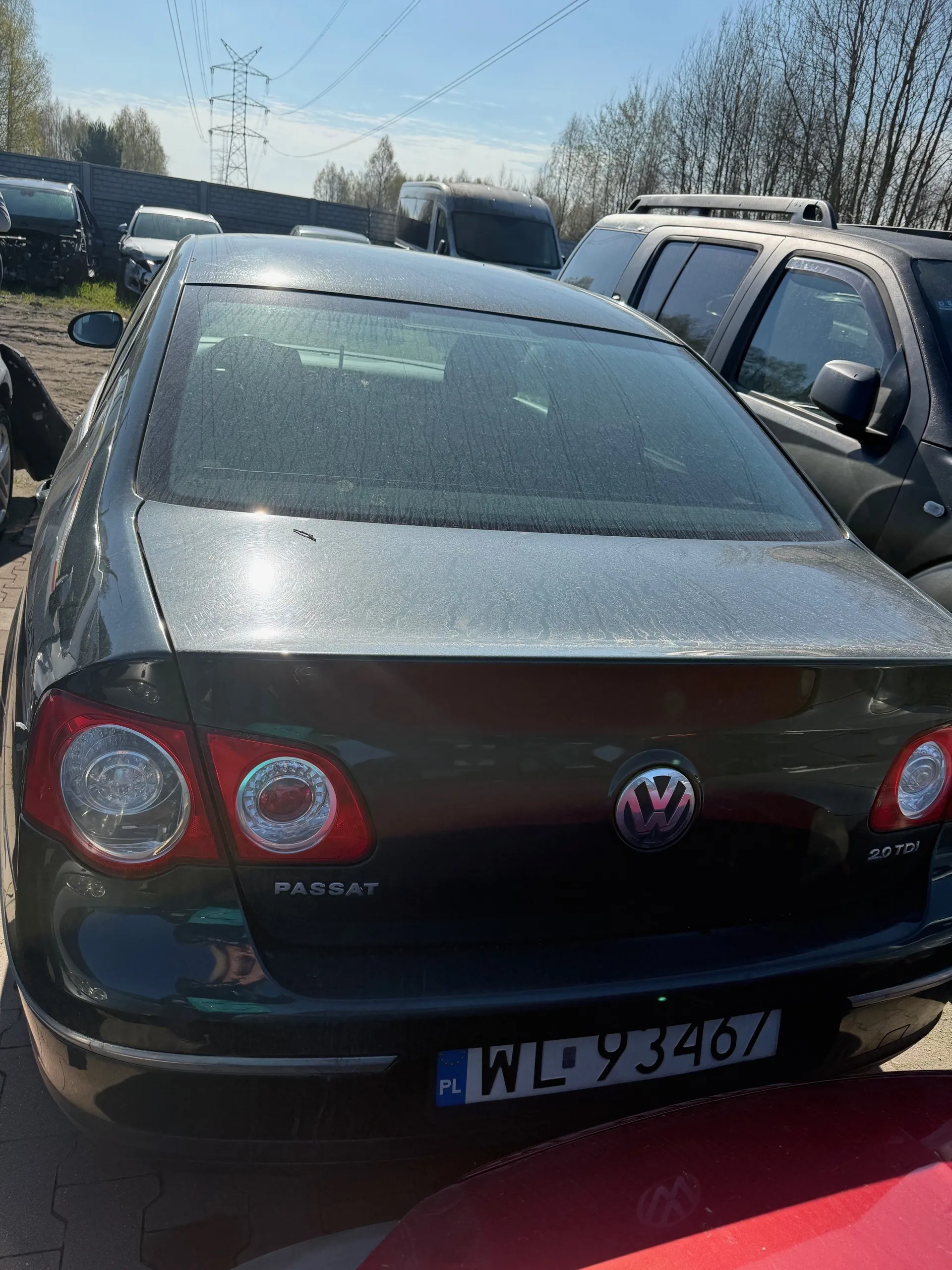 Z tyłu czarny samochód Volkswagen Passat z widocznym logo VW, napisem PASSAT oraz tablicą rejestracyjną PL WL 93467. W tle inne pojazdy, drzewa bez liści, słup energetyczny i jasne niebo. Samochód stoi na parkingowym placu z kostkami brukowymi.