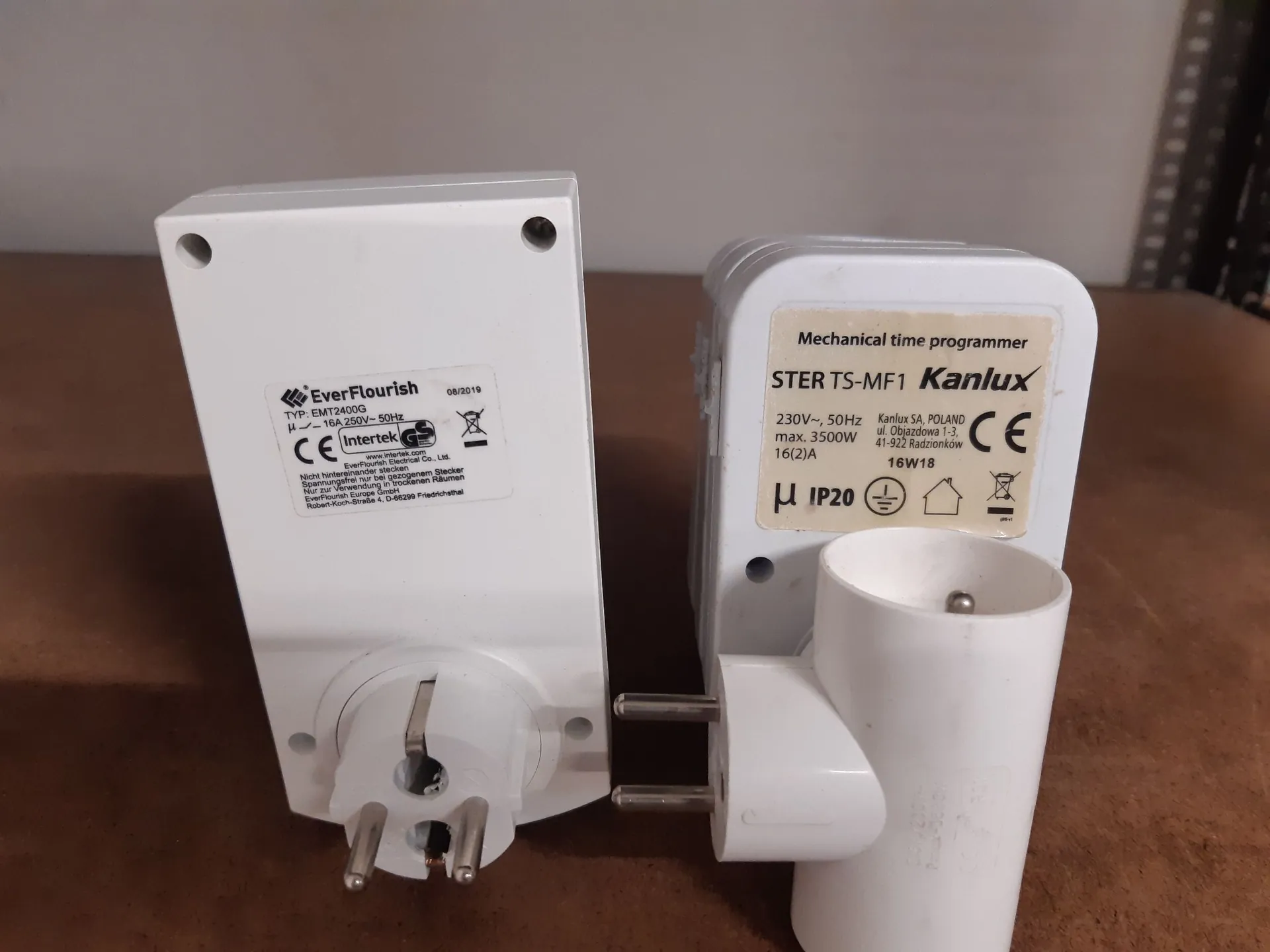 Dwa białe urządzenia elektryczne leżące obok siebie na brązowej powierzchni. Po lewej znajduje się moduł EverFlourish Typ: EMT240DG z etykietą zawierającą dane techniczne: 16A 250V~ 50Hz oraz oznaczenia CE i Intertek. Po prawej znajduje się programator czasowy Kanlux STER TS-MF1 z napisem 'Mechanical time programmer', danymi: 230V~, 50Hz, max. 3500W, 16(2)A, IP20 oraz oznaczeniem CE. Oba urządzenia posiadają wtyczki elektryczne.