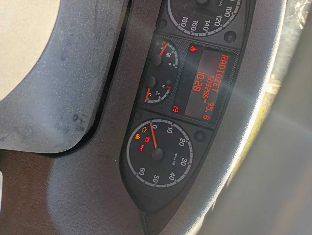 Tablica rozdzielcza samochodu z prędkościomierzem (0-180 km/h), tachometrem (0-60 x100 obr./min), wyświetlaczem centralnym pokazującym 'RADIOZET 95.6', '509286 km' oraz '10:28'. Widoczne wskaźniki paliwa (niski poziom), temperatury chłodniczki oraz lampki ostrzegawcze: pas bezpieczeństwa, akumulator, ciśnienie oleju. Fragment kierownicy widoczny w dolnej części obrazu.