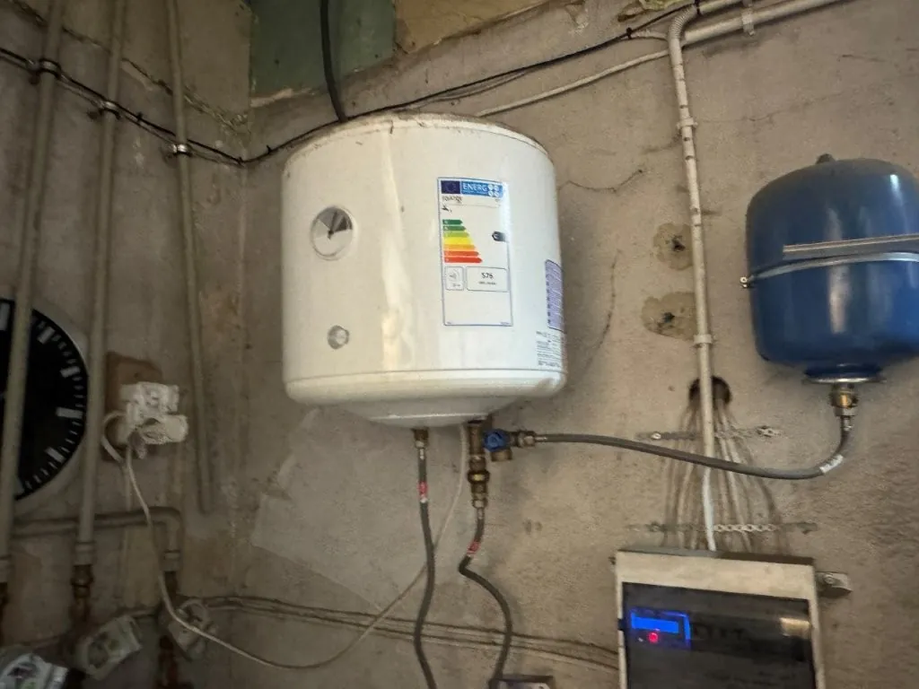 Biały, cylindryczny bojler elektryczny zamontowany na ścianie w pomieszczeniu technicznym. Na obudowie urządzenia znajduje się kolorowa etykieta energetyczna z klasami od A do G. Do bojlera podłączone są metalowe rurki i węże. Po prawej stronie widoczny jest niebieski zbiornik przeponowy, a poniżej szary panel sterowania z niebieską diodą. W tle widać rury instalacyjne biegnące wzdłuż ściany.