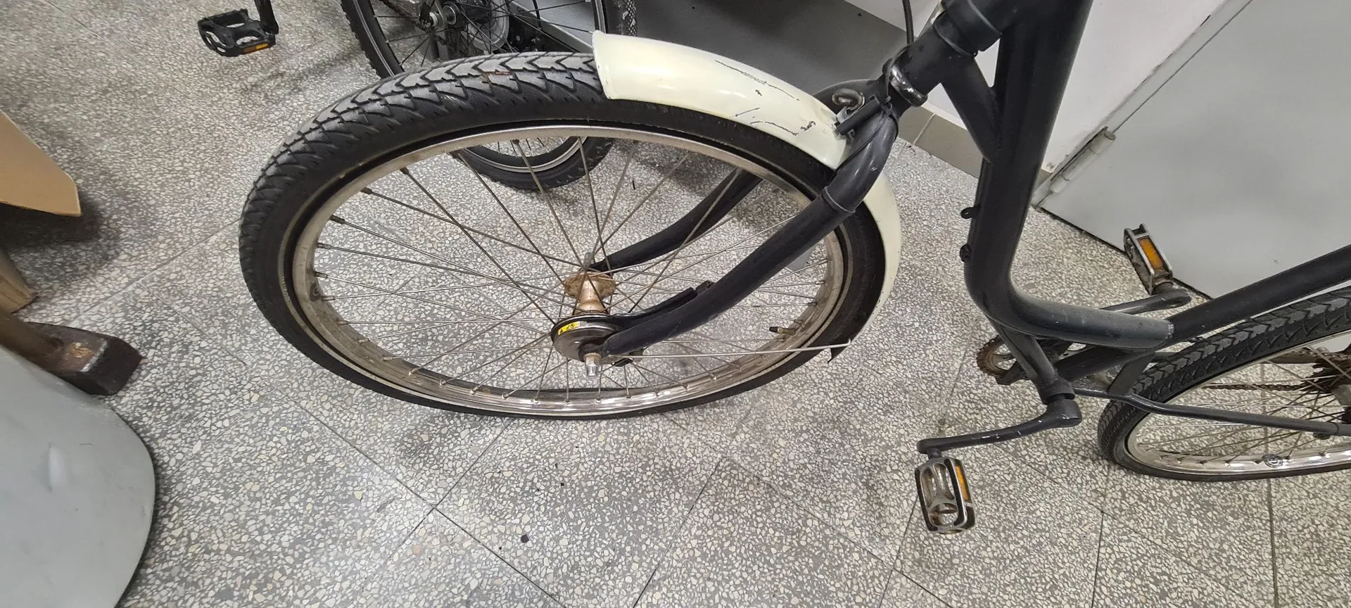Zbliżenie na przednie koło czarnego roweru z białym błotnikiem. Rower stoi na szarej, kropkowanej posadzce. Widoczna jest opona z bieżnikiem, szprychy oraz fragment ramy i pedału.