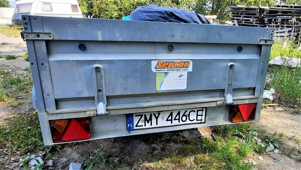 Tylny widok szarej przyczepy transportowej marki Neptun. Na tylnej klapie znajduje się logo 'Neptun' z adresem strony internetowej 'neptun-anhaenger.com' oraz mniejsza naklejka informacyjna. Na dole zamontowana jest tablica rejestracyjna o numerze ZMY 446CE z oznaczeniem kraju UE i skrótem FL. Po obu stronach znajdują się czerwone światła tylne z odblaskami. Przyczepa stoi na nierównym, trawiastym i ziemistym podłożu.