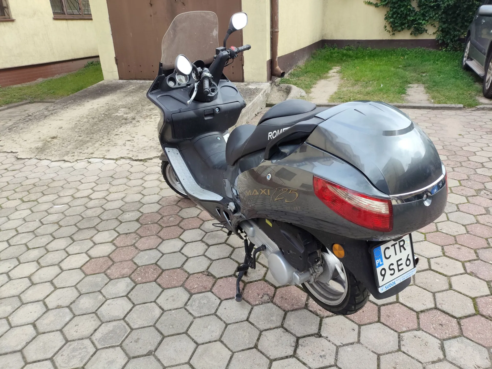 Szary skuter marki ROMA, model MAXI 125, zaparkowany na kostce brukowej o sześciokątnym kształcie. Pojazd jest widoczny z tyłu i z boku, posiada tablicę rejestracyjną o numerze CTR 9SE6 z oznaczeniem PL. W tle widać fragment jasnego budynku oraz zielony trawnik.