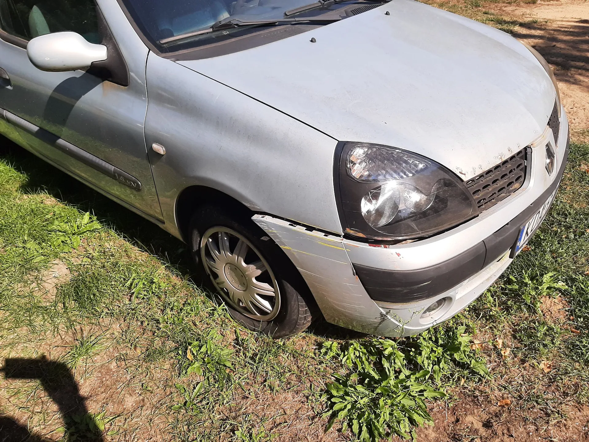 Srebrny samochód osobowy marki Renault zaparkowany na trawiastym podłożu. Widoczny jest przód pojazdu z uszkodzonym przednim zderzakiem po prawej stronie, który jest lekko odkształcony i oddzielony od nadwozia. Samochód ma ciemne reflektory i srebrne felgi aluminiowe.