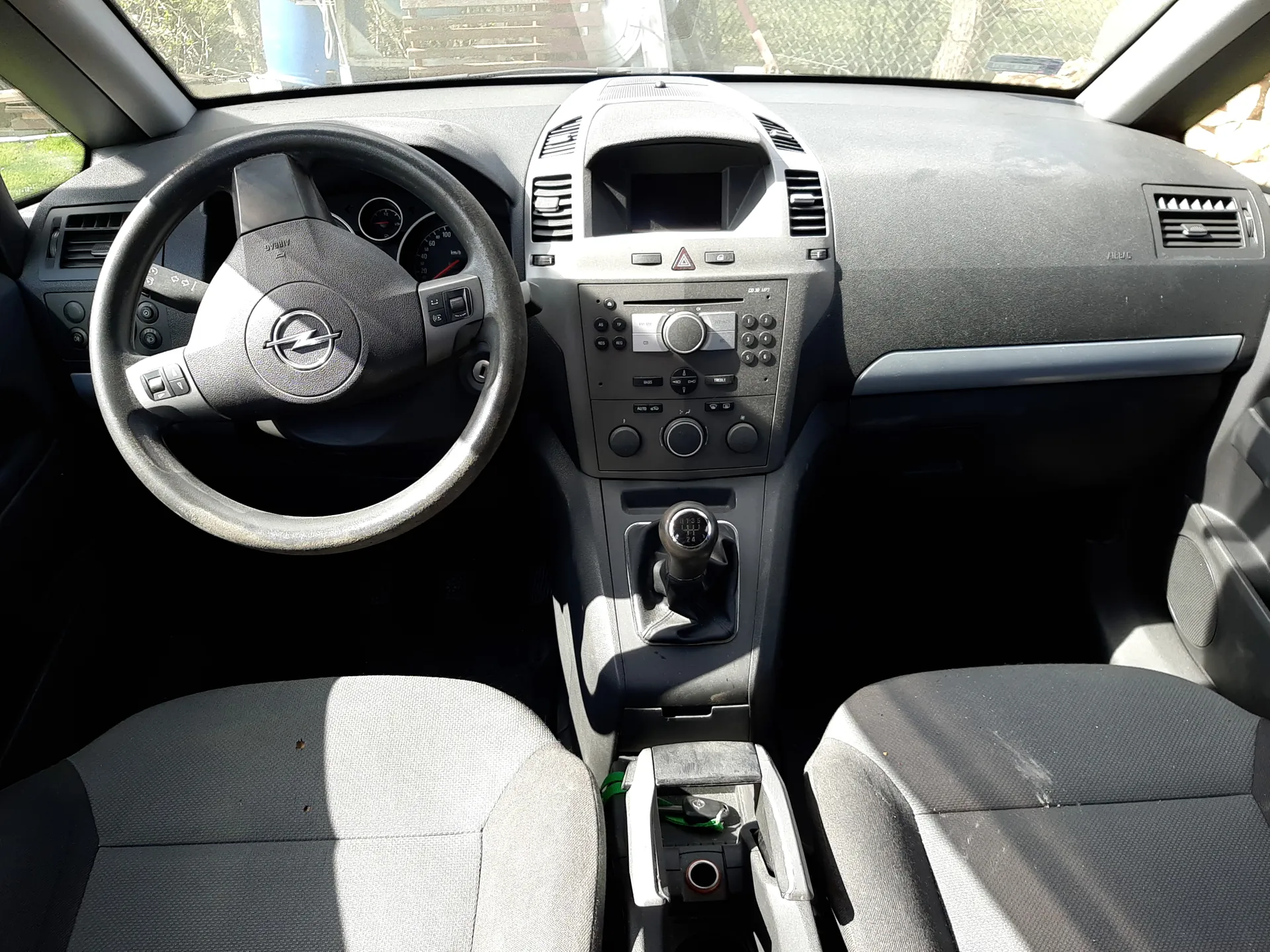 Wnętrze samochodu z widoczną kierownicą z logo Opel, deska rozdzielcza z licznikami, systemem audio i klimatyzacją, dźwignią zmiany biegów oraz przednimi siedzeniami w kolorze szaro-czarnym. Przez przednią szybę widoczny jest ogrodzenie i drzewa na zewnątrz.