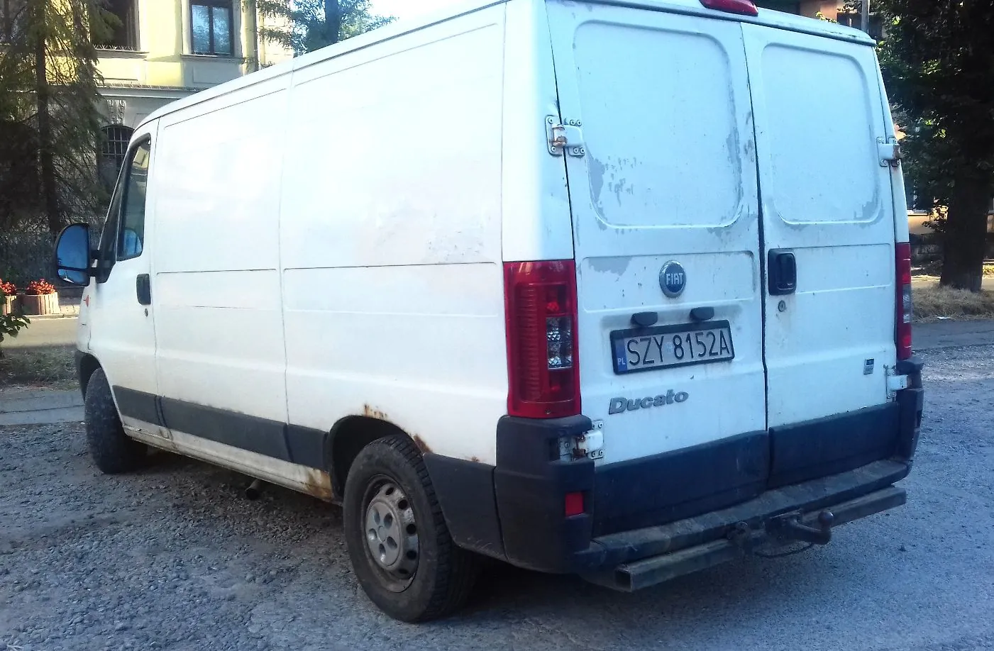 Biały furgon marki Fiat Ducato zaparkowany na żwirowym podłożu. Pojazd jest widoczny od tyłu i z lewej strony. Na tylnych drzwiach znajduje się logo Fiata oraz polska tablica rejestracyjna o numerze SZY 8152A. Nad tablicą widnieje napis Ducato. Samochód posiada widoczne ślady rdzy na dolnej części nadwozia oraz zabrudzenia.