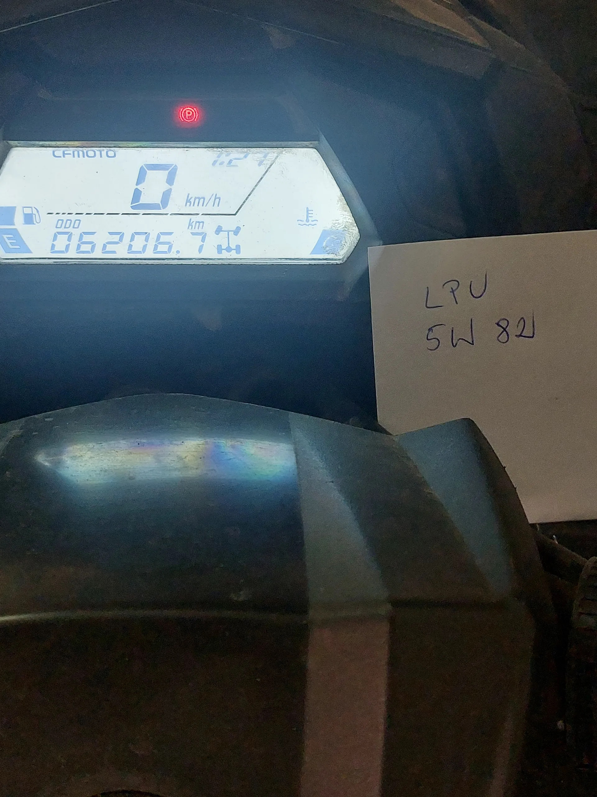 Zbliżenie na cyfrowy zegar samochodowy lub motocyklowy marki CFMOTO. Wyświetlacz pokazuje prędkość 0 km/h, przebieg 06206.7 km oraz czerwone światło ostrzegawcze. Nad wyświetlaczem znajduje się biały kartonik z ręcznie napisanym tekstem: LRU 5W 82J. Tło stanowi ciemna, metalowa powierzchnia pojazdu.