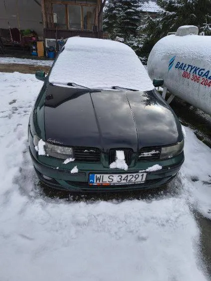 Zdjęcie przedstawia ciemnozielony samochód osobowy pokryty śniegiem, stojący na śnieżnej powierzchni. Na masce widoczny jest napis 'Audi'. Tablica rejestracyjna oznaczona jako 'WLS 34291' z niebieskim pasem po lewej stronie. Po prawej stronie samochodu znajduje się biały zbiornik gazowy z napisem 'BALTYSKAGAZ' oraz numerem telefonu '800 306 204'. W tle widoczny jest dom z oknami i przedmiotami na zewnątrz, a także drzewa pokryte śniegiem. Całość otoczona jest śniegiem.
