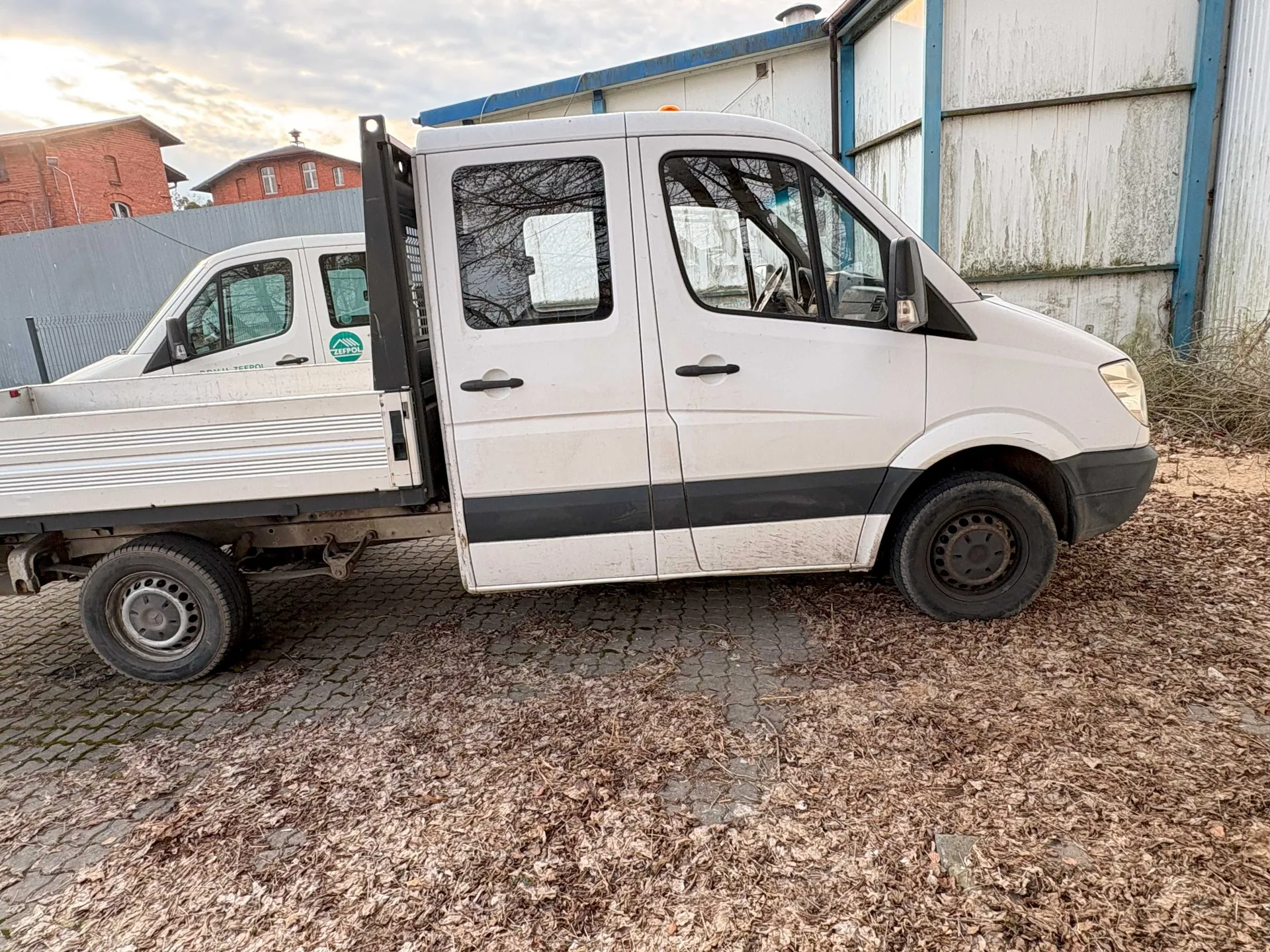 Biały samochód dostawczy typu pickup z czarną listwą po bokach, zaparkowany na brukowanej powierzchni pokrytej suchymi liśćmi. Po lewej stronie widoczna kabina z dwoma drzwiami i napisem 'ZEPOL' w zielonym okręgu na boku. Tło stanowią budynek z białymi i niebieskimi ścianami, czerwone budynki z cegły oraz ogrodzenie. Niebo częściowo zachmurzone.