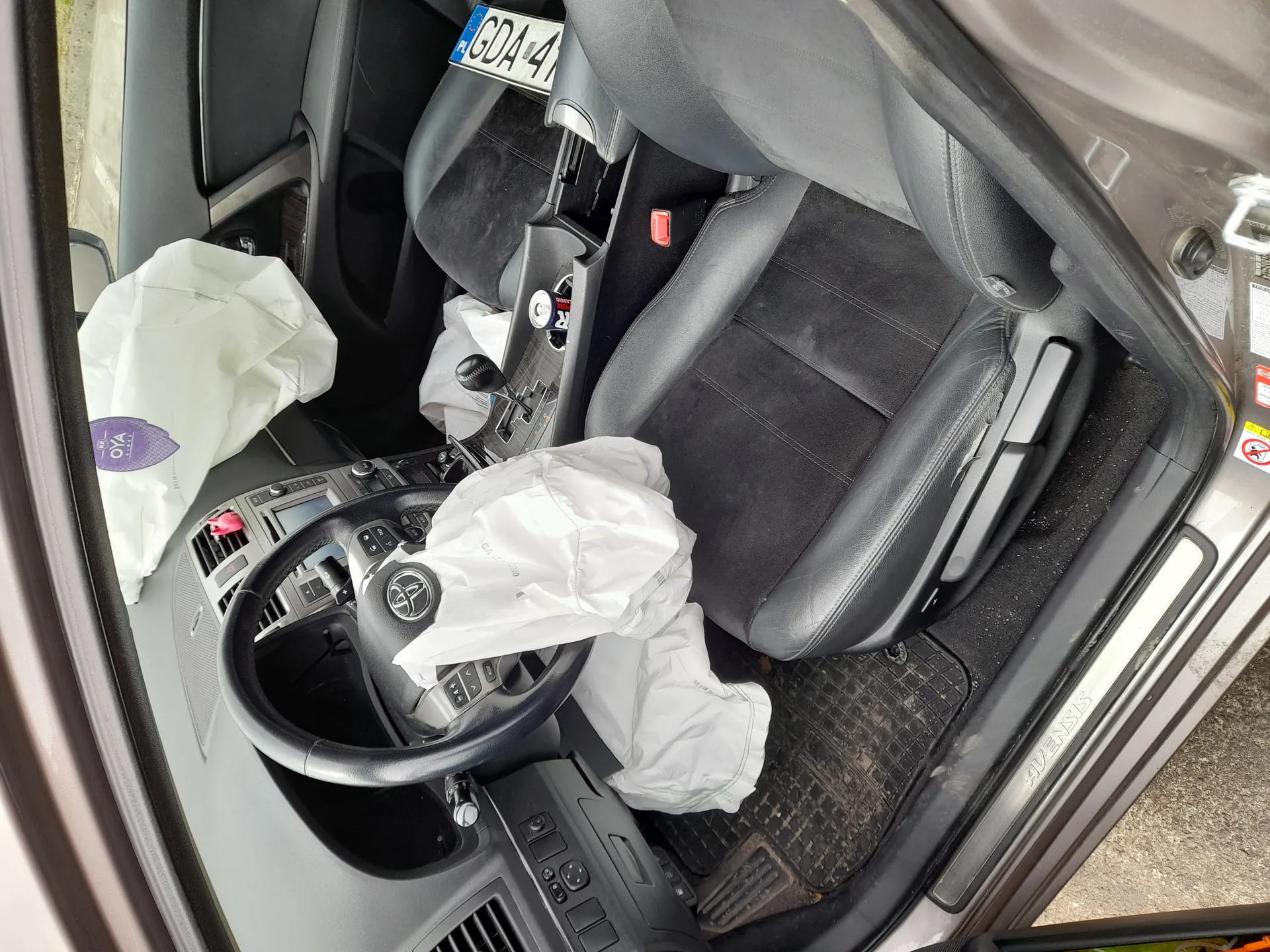 Wnętrze samochodu marki Toyota Avensis z rozwiniętymi poduszkami powietrznymi. Widoczne czarne skórzane fotele, kierownica z logo Toyoty, deska rozdzielcza z przyciskami, dźwignia biegów oraz tablica rejestracyjna z napisem GDA 4. Na podłodze leżą białe worki, a na drzwiach widoczny napis AVENSIS oraz znak zakazu palenia. Wnętrze wydaje się nieporządne, z brudnymi dywanikami.
