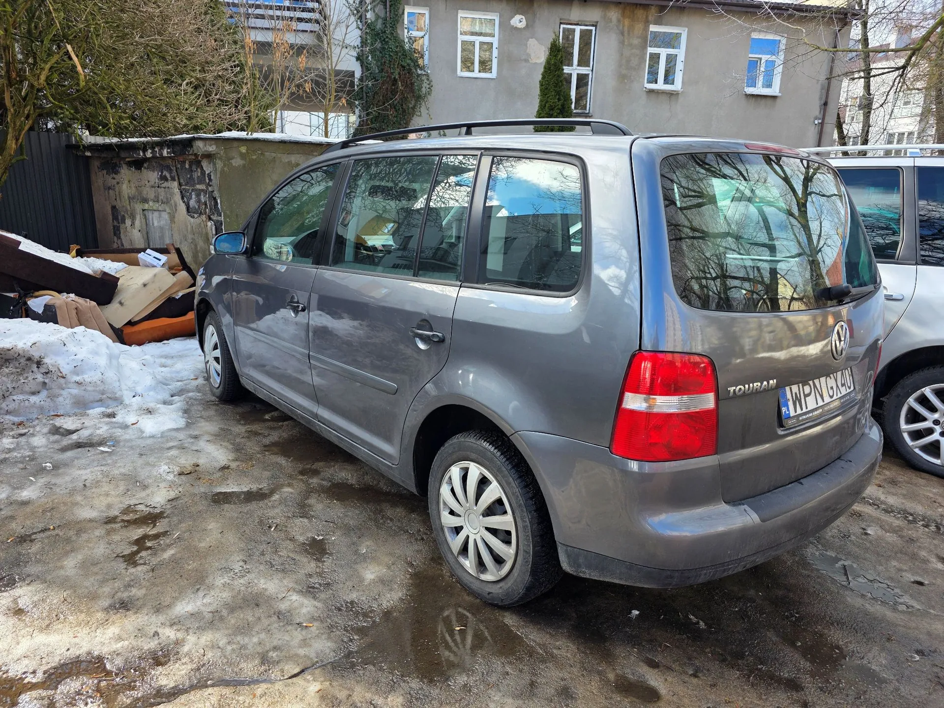 Szary samochód marki Volkswagen Touran zaparkowany na błotnistym podłożu z resztkami śniegu. Pojazd jest widoczny z tyłu i z boku. Na klapie bagażnika znajduje się napis 'TOURAN' oraz tablica rejestracyjna o numerze 'WPN GX40'. W tle widoczny jest stary budynek z oknami oraz sterta materiałów budowlanych i gruzu po lewej stronie.