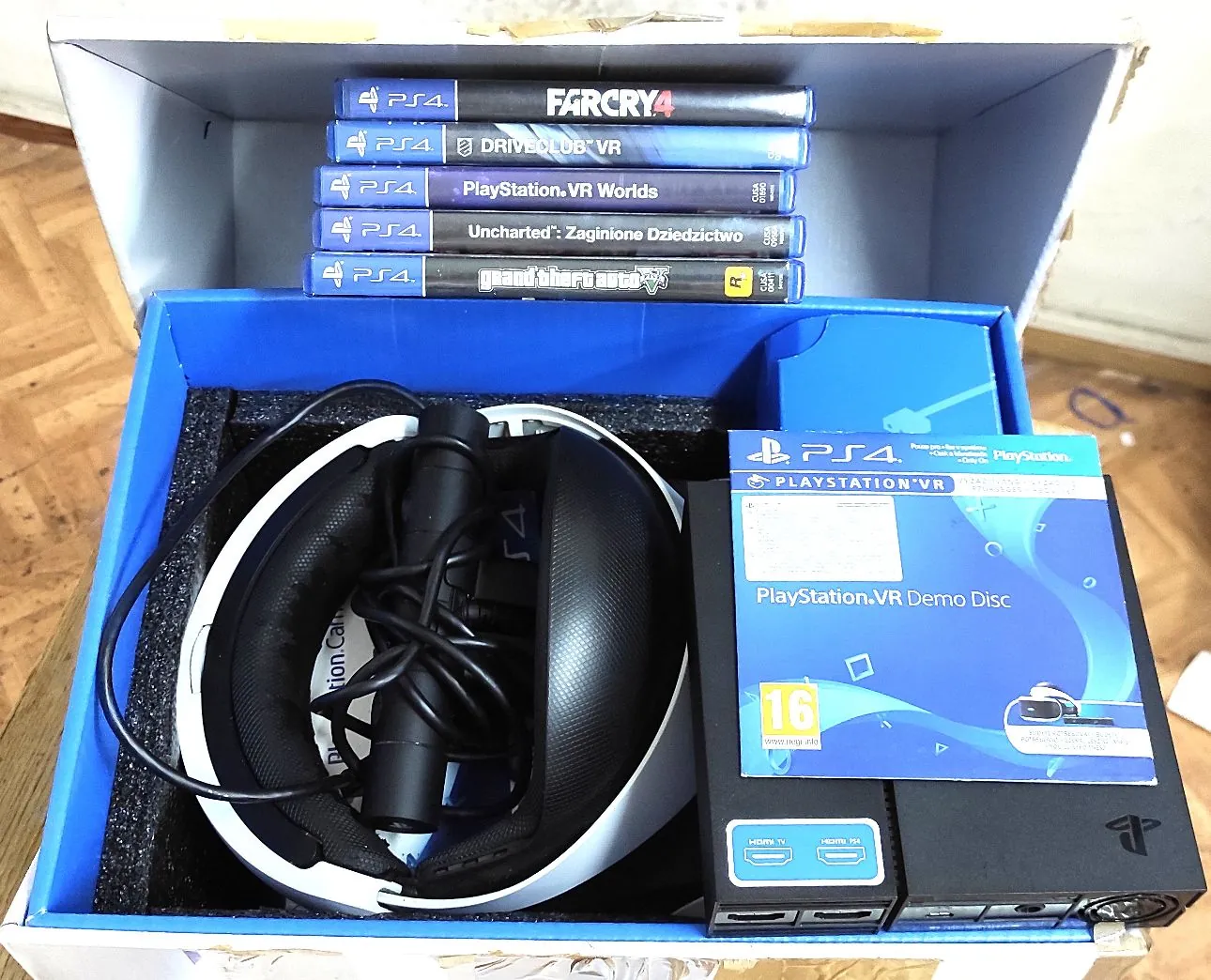 Otwarta niebieska skrzynka zawierająca zestaw PlayStation VR oraz gry na konsolę PS4. W górnej części, na wewnętrznej klapie pudełka, ułożone jest pięć gier: 'Far Cry 4', 'Driveclub VR', 'PlayStation VR Worlds', 'Uncharted: Zaginione Dziedzictwo' oraz 'Grand Theft Auto V'. Wewnątrz pudełka znajduje się gogle VR z czarnymi gąbkami i dołączonym kablem, a obok nich pudełko z płytą 'PlayStation VR Demo Disc' z oznaczeniem PEGI 16 oraz czarne urządzenie procesora VR z portami HDMI i USB.
