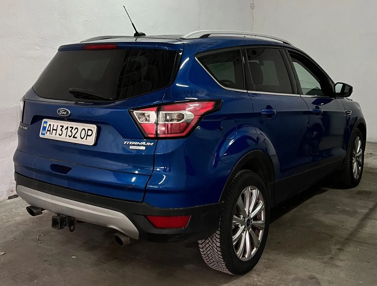 Niebieski samochód osobowy typu SUV marki Ford, widok z tyłu i boku. Na tylnym zderzaku widoczny napis "TITANIUM", logo Ford oraz tablica rejestracyjna z numerem "AH 3132 OP" z flagą Ukrainy po lewej stronie. Samochód stoi w pomieszczeniu z betonową podłogą i białą ścianą. Widoczne są czarne felgi, czerwone światła tylnego oraz dachowy antena.