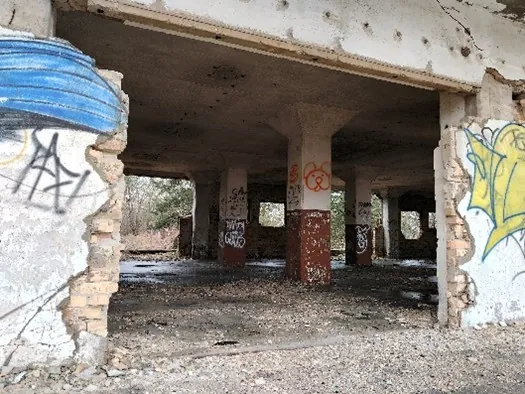 Wnętrze zniszczonego, betonowego budynku z widocznymi filarami podtrzymującymi strop. Ściany i kolumny są pokryte graffiti oraz odpryskami tynku, odsłaniając cegły. Podłoga jest brudna, pokryta gruzem i pyłem. W tle widoczne są otwory okienne prowadzące na zewnątrz, gdzie widać bezlistne drzewa.