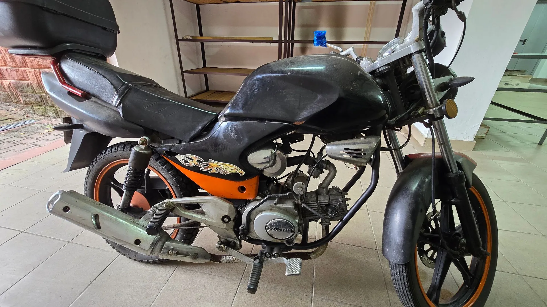 Czarny motocykl zaparkowany w pomieszczeniu na jasnej, kafelkowej podłodze. Pojazd posiada czarny bak, czarne siedzenie oraz pomarańczowe detale na felgach i owiewkach. Widoczny jest srebrny wydech, silnik oraz przedni i tylny błotnik. W tle znajdują się drewniane regały i biała ściana.