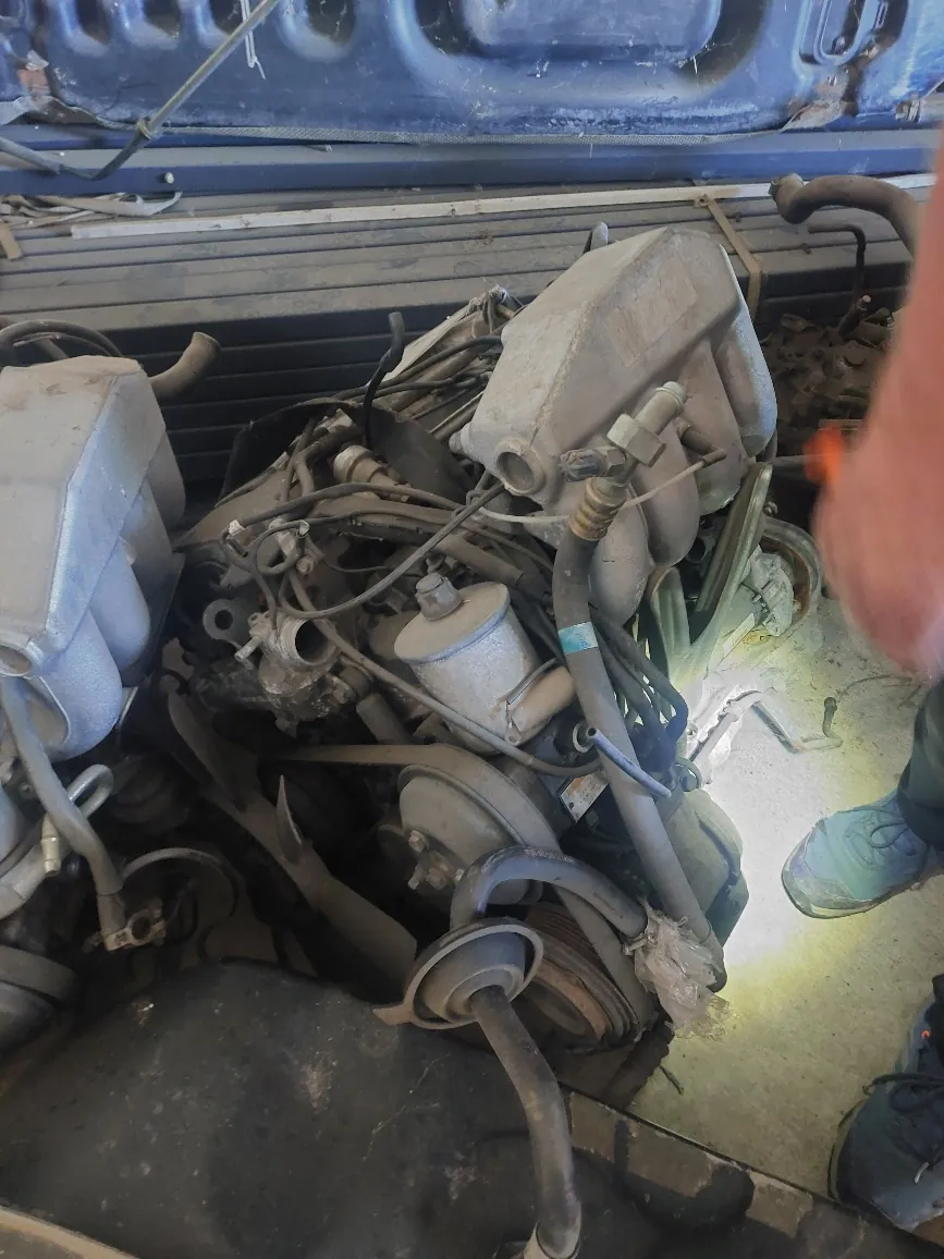 Zbliżenie na zakurzony silnik spalinowy w komorze silnika pojazdu. Widoczne są elementy mechaniczne, przewody, zbiornik wyrównawczy oraz paski napędowe. W prawym dolnym rogu widoczna jest część nogi osoby w niebieskim obuwiu.