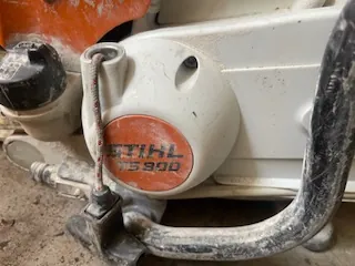 Zbliżenie na biały element mechaniczny z pomarańczową etykietą z napisem STIHL 75900. Widoczne są metalowe części, w tym hak i czarna nakrętka. Tło stanowi betonowa powierzchnia. Elementy są umieszczone w naturalnej orientacji.