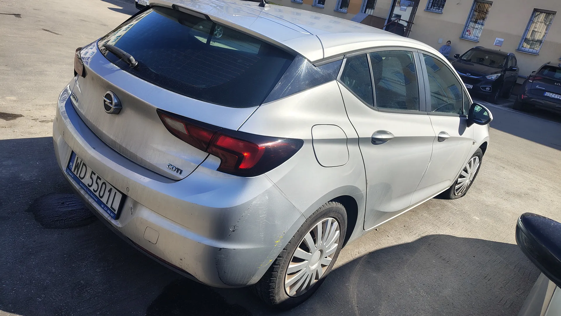 Srebrny samochód Opel Corsa z tyłu, widoczny znak rejestracyjny PL WD 5501L oraz napis CDTi na bagażniku. Samochód zaparkowany na asfaltowym placu parkingowym, w tle budynki z oknami, czarny samochód Hyundai i osoba stojąca blisko wejścia. Na tylnej szybie widoczny wycieraczka, a na masce reflektory i logo marki.