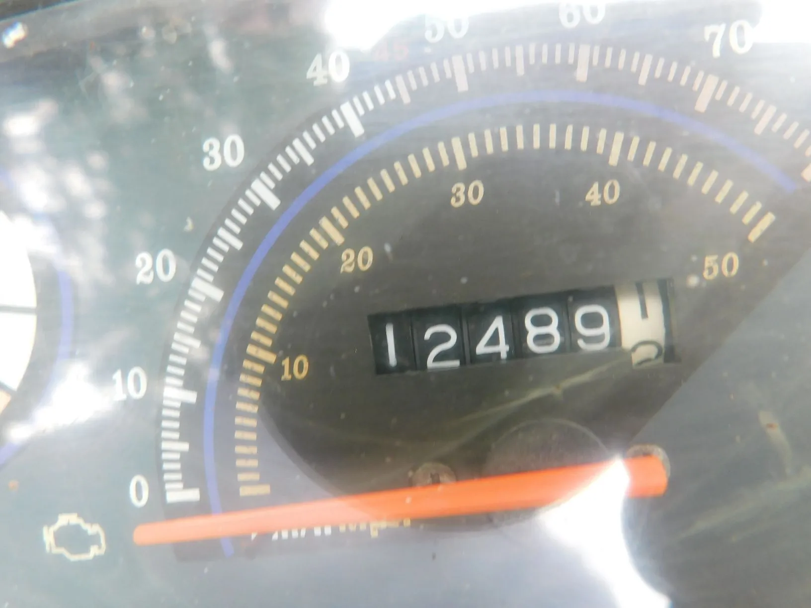 Zbliżenie na licznik przebiegu w samochodzie. Widoczny jest analogowy licznik z cyframi 12489 oraz skala prędkościomierza z oznaczeniami od 0 do 70. Na dole znajduje się pomarańczowa wskazówka oraz ikona kontrolki silnika.