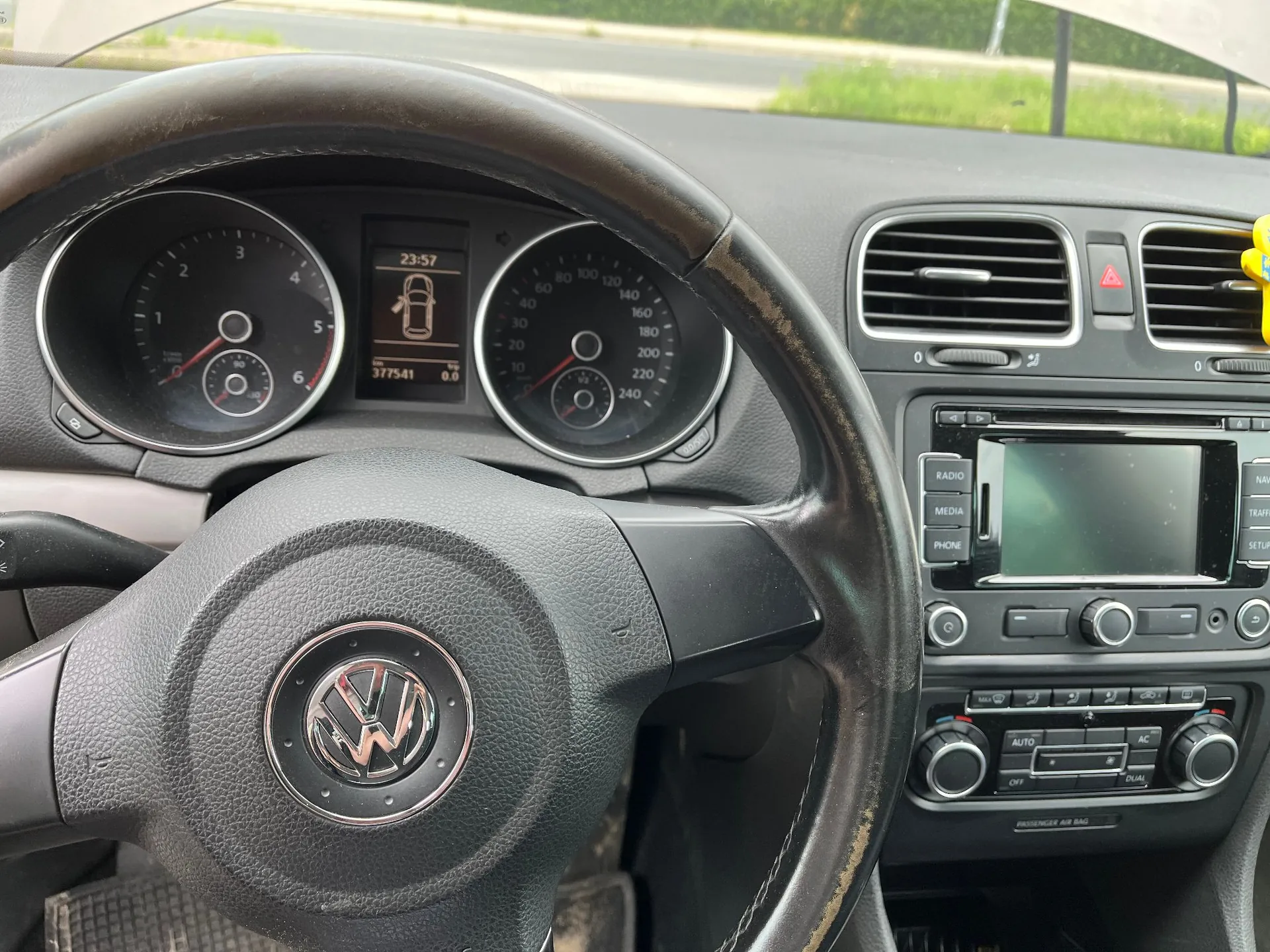 Wnętrze samochodu marki Volkswagen widziane z perspektywy kierowcy. Widoczna jest czarna kierownica z logo VW w centrum, deska rozdzielcza z dwoma analogowymi wskaźnikami prędkości i obrotów, centralny wyświetlacz pokazujący godzinę 23:57 i przebieg 377541 km oraz konsola środkowa z radiem i panelem klimatyzacji.