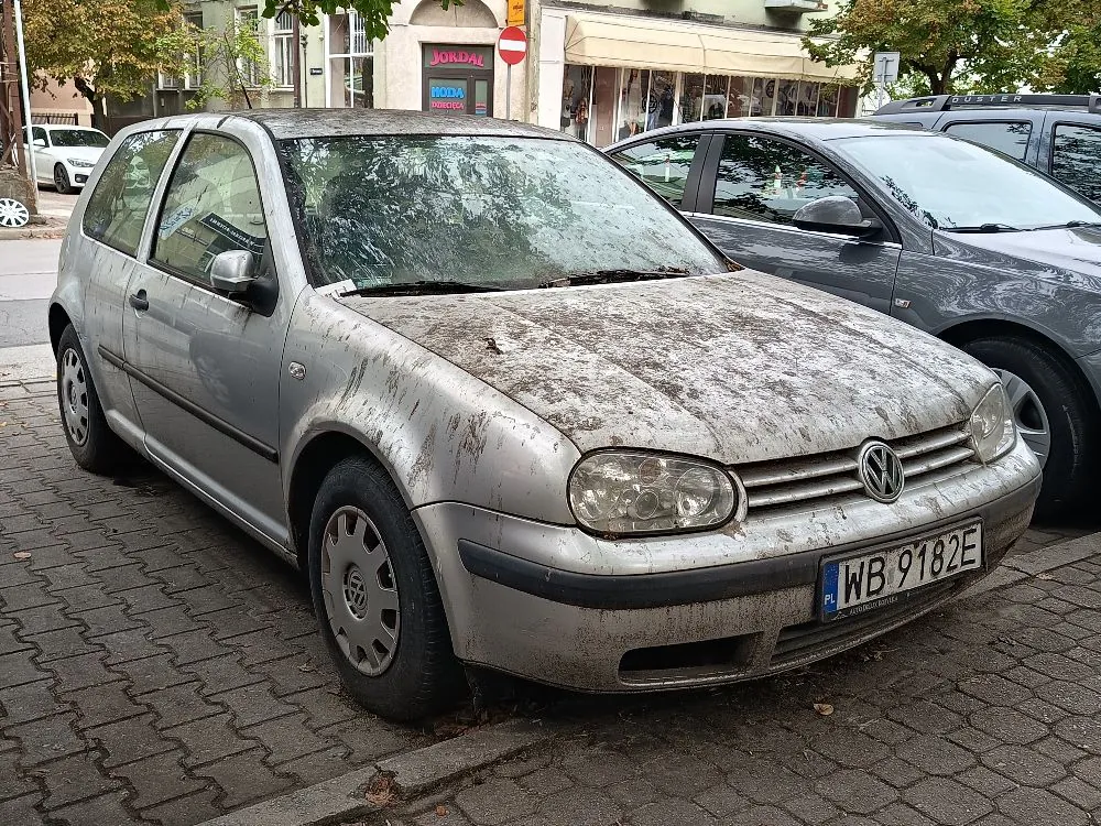 Srebrny samochód marki Volkswagen Golf zaparkowany na kostce brukowej. Pojazd jest bardzo brudny, pokryty warstwą błota i pyłu na masce, dachu oraz przedniej szybie. Samochód posiada polską tablicę rejestracyjną o numerze WB 9182E. W tle widoczne są inne zaparkowane auta, budynki oraz drzewa.