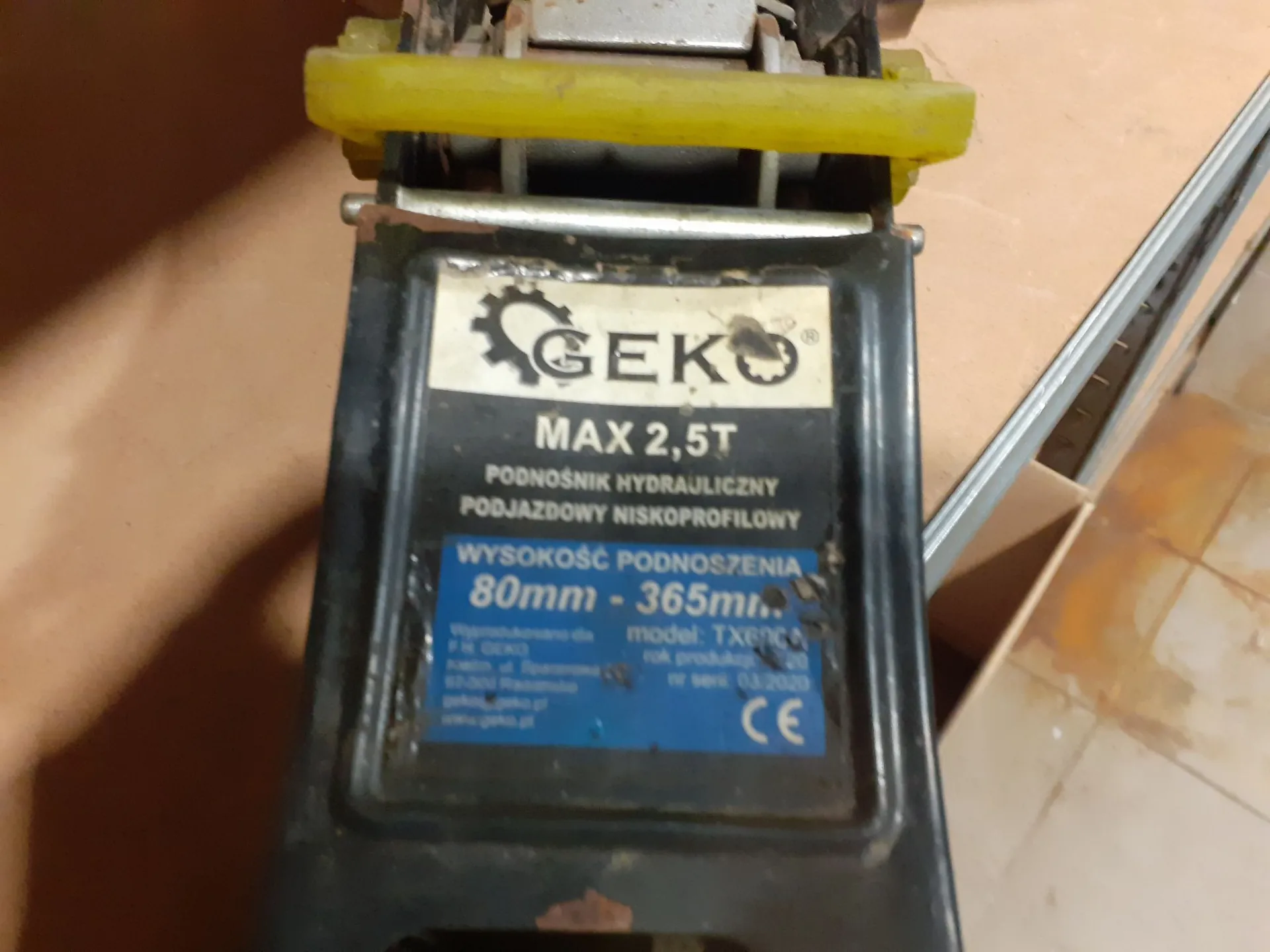 Zbliżenie na tabliczkę znamionową czarnego podnośnika hydraulicznego. Na białym tle widnieje logo 'GEKO' z symbolem koła zębatego, a poniżej tekst: 'MAX 2,5T', 'PODNOŚNIK HYDRAULICZNY PODJAZDOWY NISKOPROFILOWY'. Na niebieskim tle znajduje się napis 'WYSOKOŚĆ PODNOSZENIA 80mm - 365mm', a pod nim mniejszym drukiem informacje o modelu TX60NC, roku produkcji 03/2020 oraz znak CE.