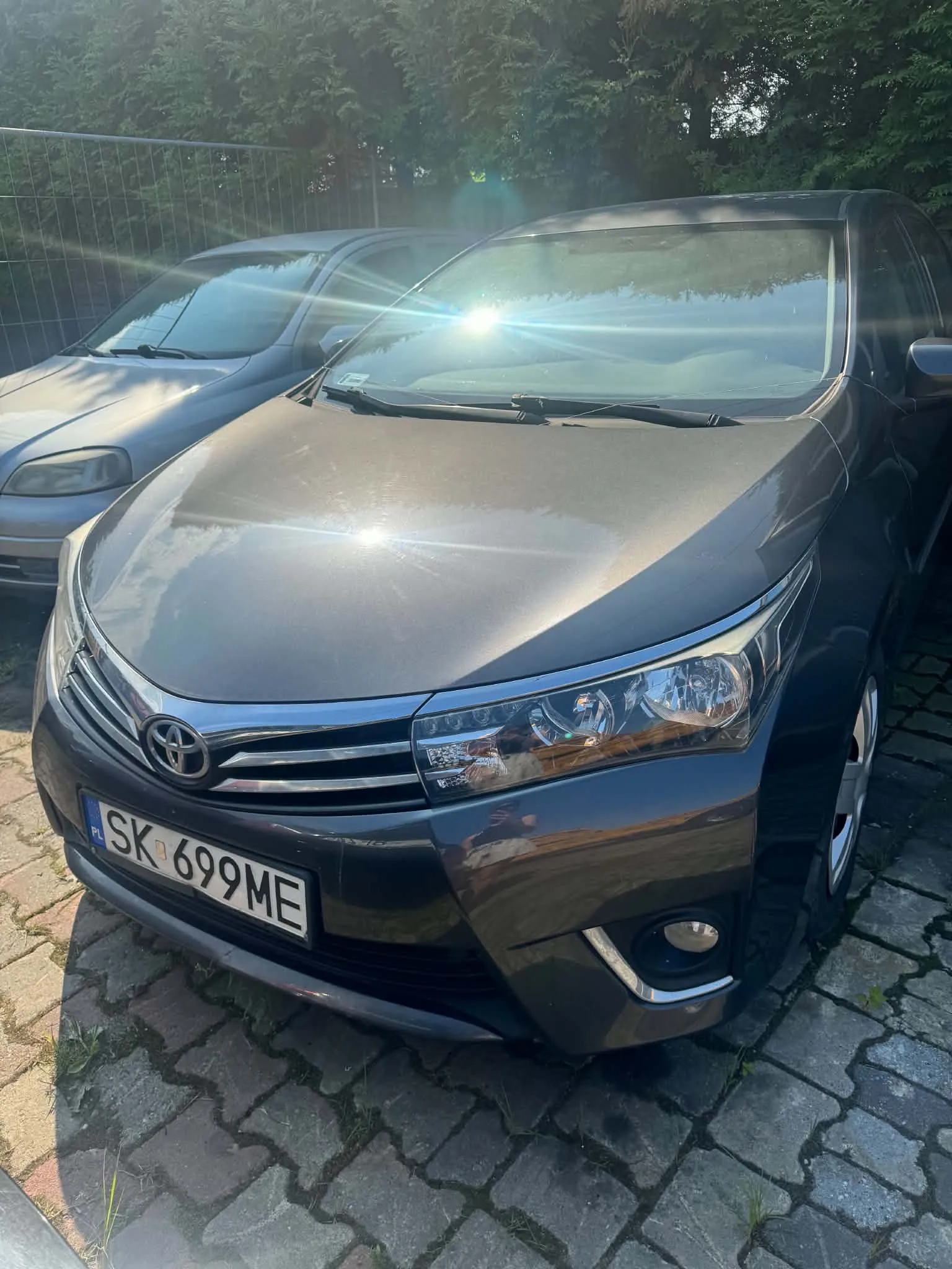 Zdjęcie przodu ciemnoszarego samochodu marki Toyota zaparkowanego na kostce brukowej. Samochód ma polską tablicę rejestracyjną o numerze SK 699ME. Obok stoi inny, starszy samochód w kolorze szarym. W tle widoczne są zielone drzewa i ogrodzenie z siatki.