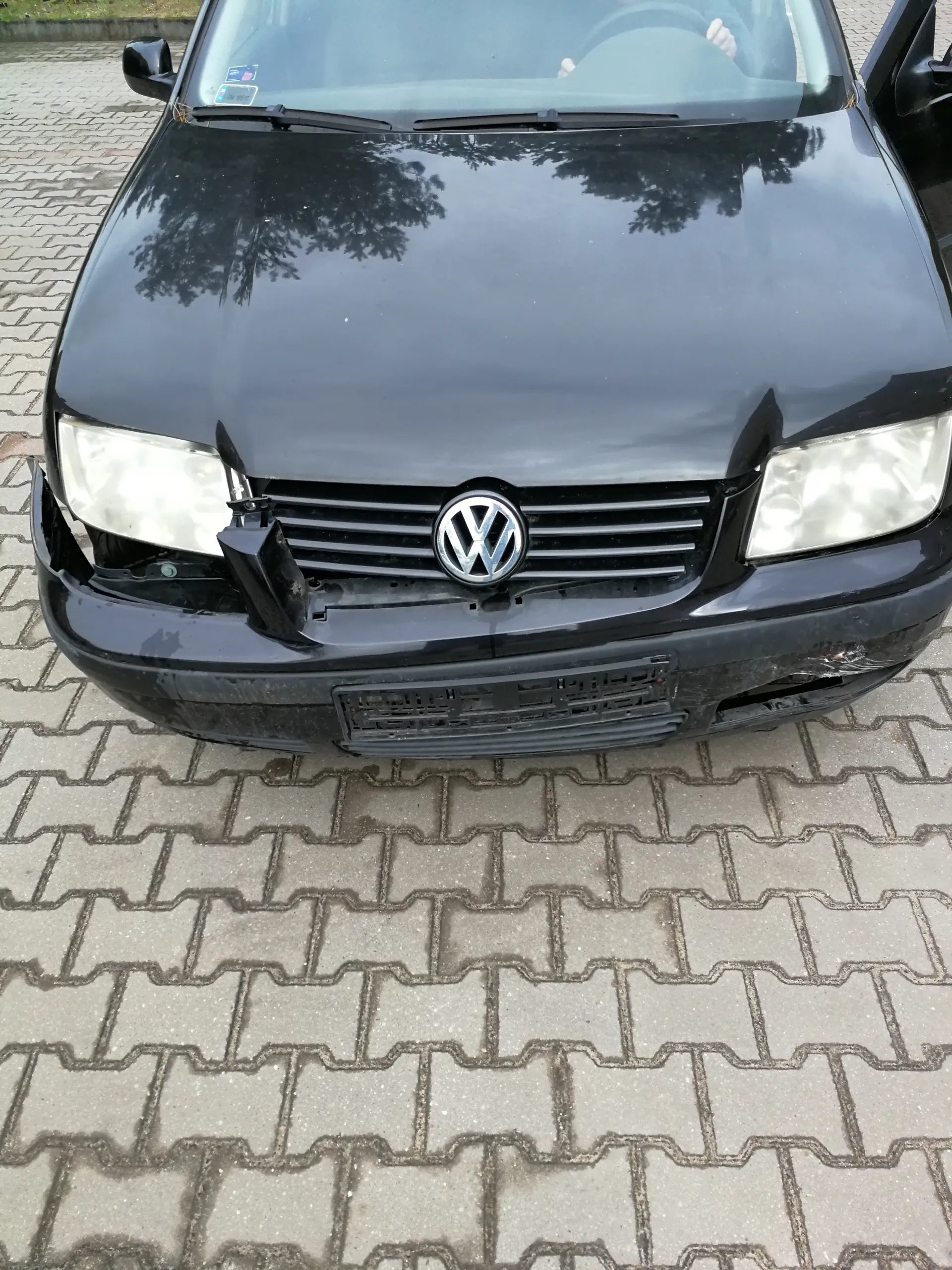Czarny samochód marki Volkswagen z widocznym logo na atrapie chłodnicy, włączonymi reflektorami i uszkodzonym lewym przednim zderzakiem. Samochód zaparkowany na brukowanej powierzchni. Na szybie przedniej widoczne są małe naklejki, a w odbiciu na masce drzewa. Tablica rejestracyjna jest pusta.