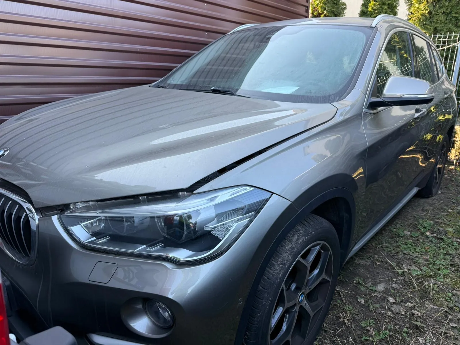 Srebrny SUV marki BMW zaparkowany na podwórku. Widok z przodu i boku. Tło stanowi brązowa metalowa brama garażowa oraz zielone krzewy i ogrodzenie. Samochód ma czarne felgi i reflektory. Na ziemi widoczna trawa i sucha trawa.