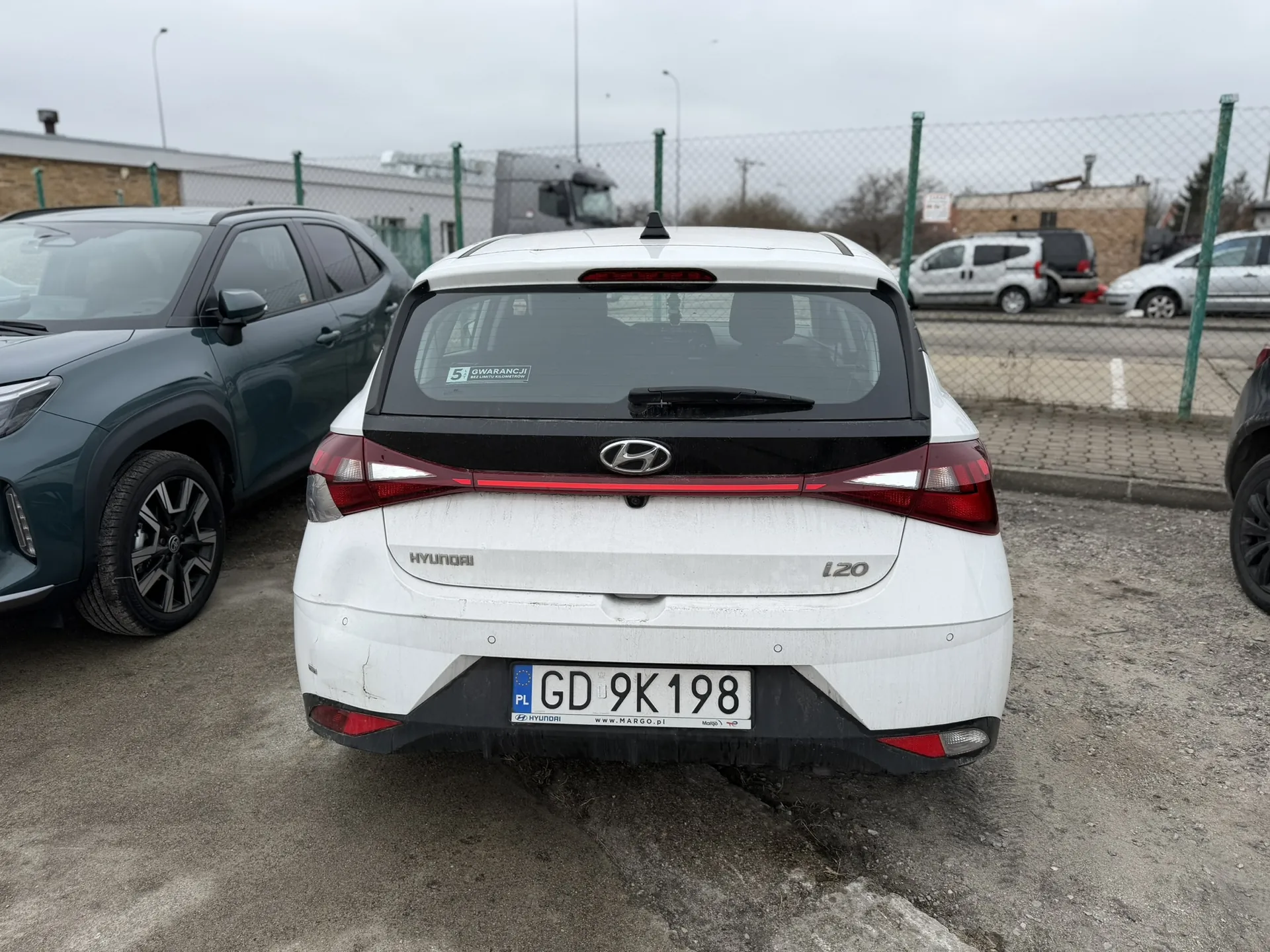 Biały samochód Hyundai i20 z tyłu, z tablicą rejestracyjną PL GD 9K198. Na szybie tylnej naklejka z napisem 5 GWARANCJI. W tle parking z innymi pojazdami, ogrodzenie siatkowe, budynki i pochmurne niebo. Po lewej stronie ciemnozielony SUV, po prawej fragment czarnego samochodu.