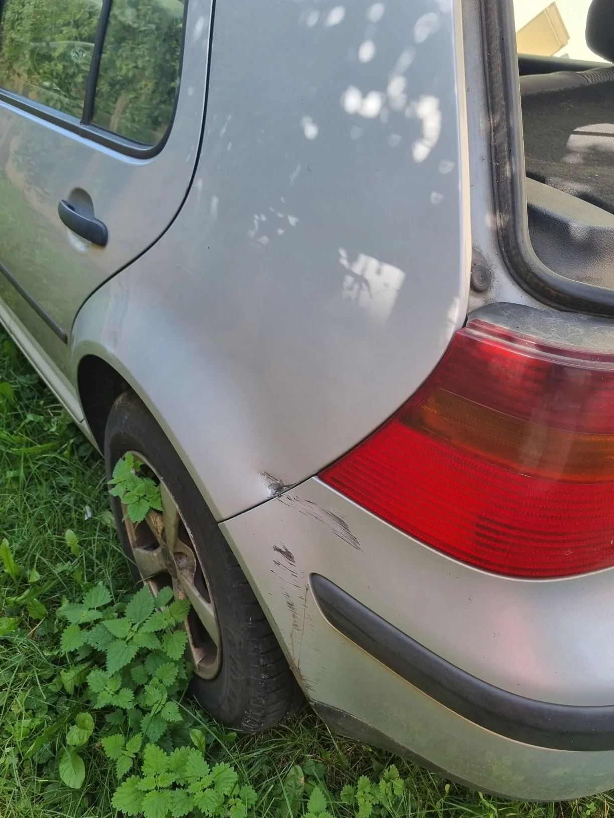 Zbliżenie na tylny prawy błotnik i lampę srebrnego samochodu. Na lakierze błotnika widoczne są zarysowania i wgniecenie w pobliżu tylnej lampy. Samochód stoi na trawie, która częściowo zasłania tylne koło.