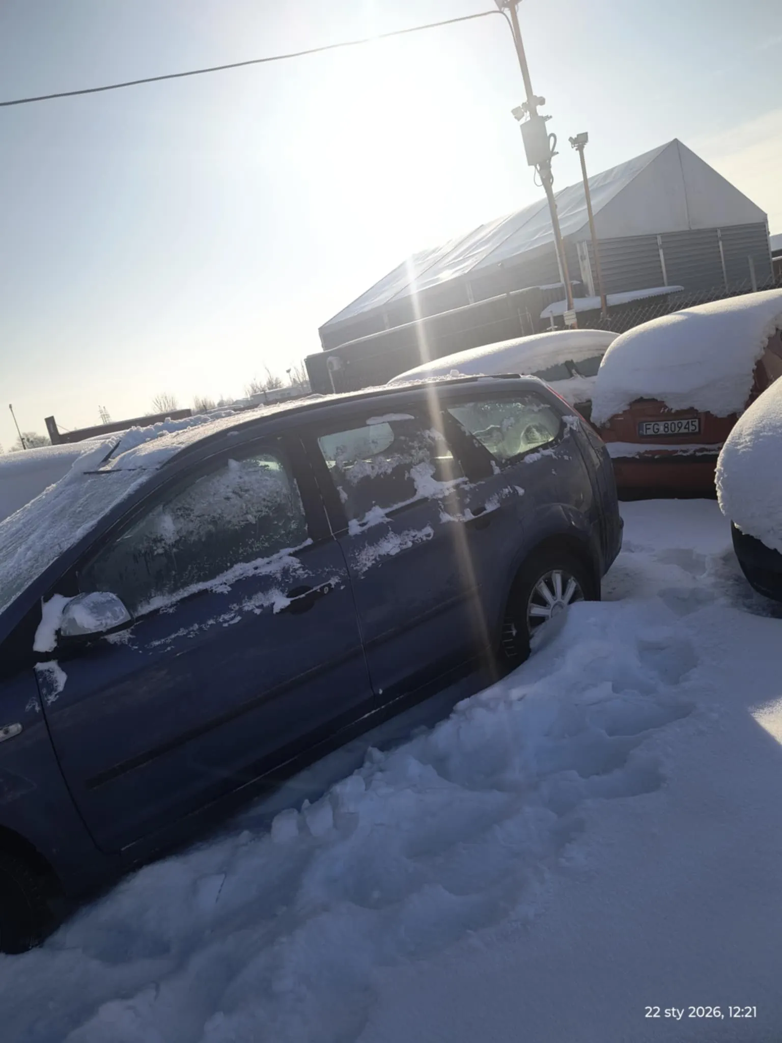 Zdjęcie przedstawia parking pokryty śniegiem z kilkoma samochodami. W pierwszym planie znajduje się niebieski samochód z widocznymi śladami śniegu na szybach i nadwoziu. Za nim stoi czerwony samochód z tablicą rejestracyjną FG 80945, również pokryty śniegiem. W tle widoczny jest budynek przemysłowy z białym dachem oraz słupy energetyczne. Słońce świeci jasno, tworząc poświatę. W prawym dolnym rogu znajduje się znacznik czasu: 22 sty 2026, 12:21. Na śniegu widoczne są ślady stóp.
