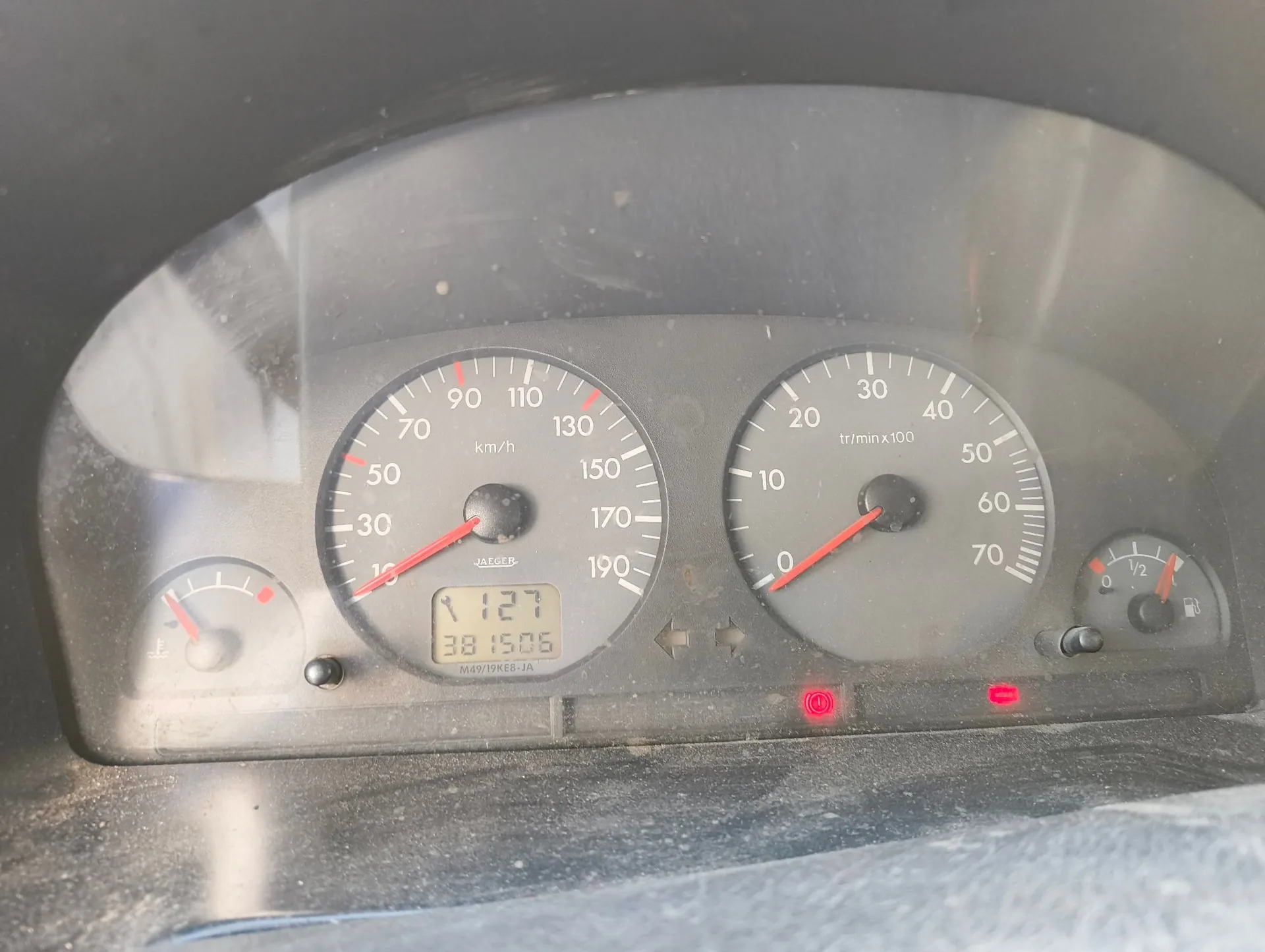 Zbliżenie na deskę rozdzielczą samochodu. Widoczny jest prędkościomierz z maksymalną wartością 190 km/h, obrotomierz z maksymalną wartością 70 (x100 tr/min) oraz wskaźnik poziomu paliwa. Poniżej prędkościomierza znajduje się cyfrowy wyświetlacz z liczbami 127 oraz 38150.5. W dolnej części prędkościomierza widnieje napis JAEGER oraz kod M49/19KE8-JA. Na desce zapalone są czerwone kontrolki.