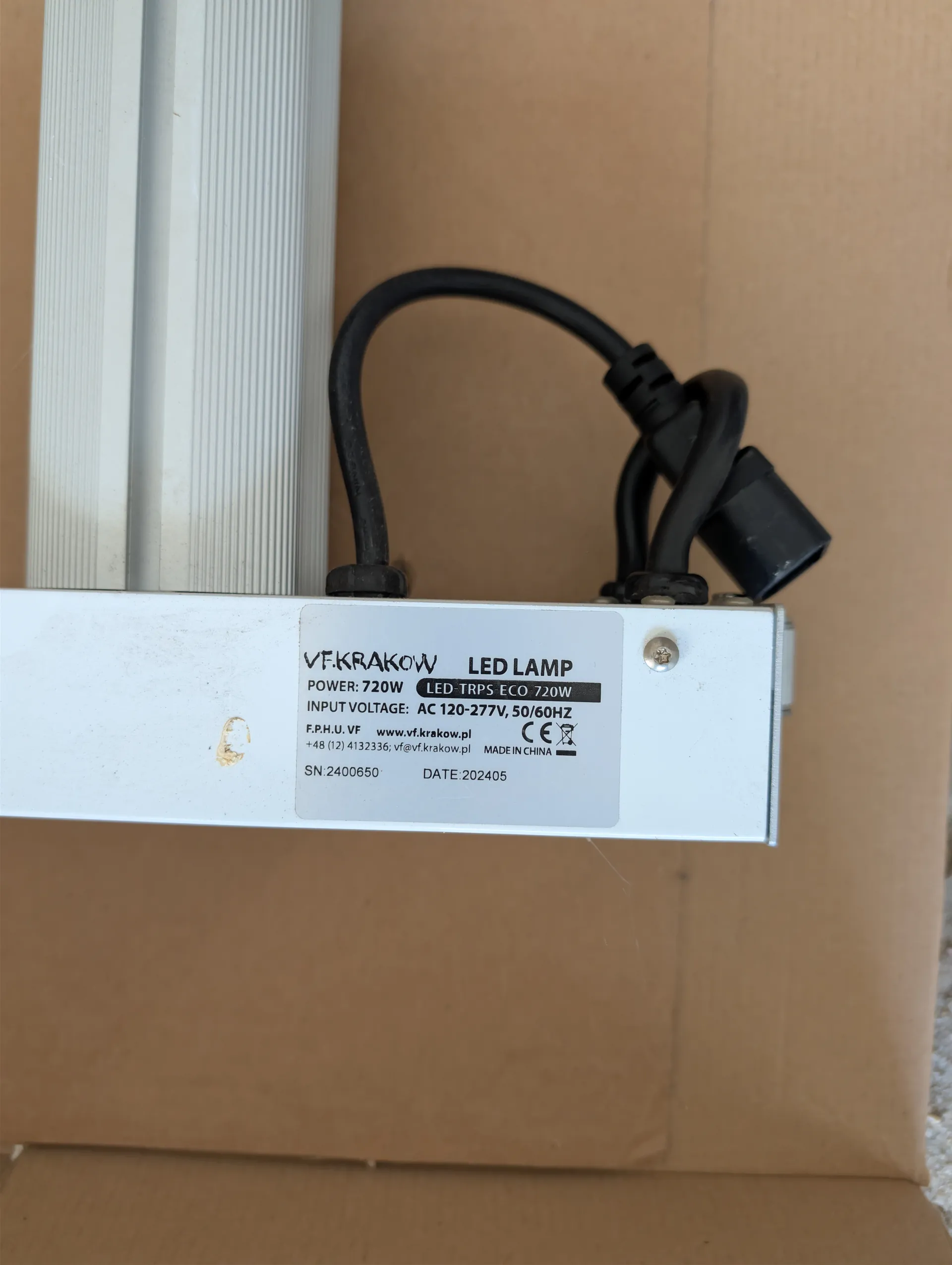 Biały LED LAMP marki VF.KRAKOW z czarnym kablem zasilającym. Na białej etykiecie widoczny tekst: VF.KRAKOW LED LAMP, POWER: 720W, LED-TRPS ECO 720W, INPUT VOLTAGE: AC 120-277V, 50/60HZ, F.P.H.U. VF www.vf.krakow.pl, +48 (12) 4132336; vf@vf.krakow.pl, MADE IN CHINA, SN:2400650, DATE:202405. Obok etykiety znajduje się mały brązowy plamka. Tło stanowi brązowe kartonowe pudełko.