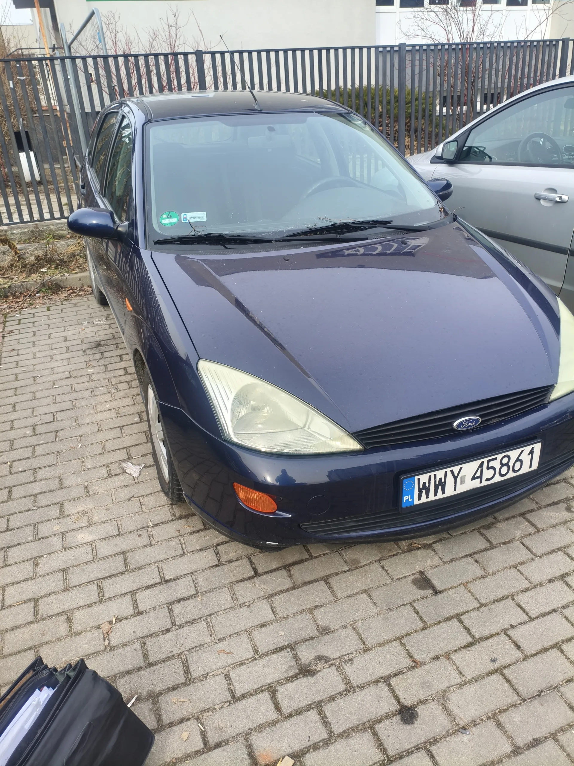 Niebieski samochód marki Ford zaparkowany na brukowanej powierzchni. Widoczna tablica rejestracyjna WWY 45861 z kodem PL oraz zielony znaczek na przedniej szybie. W tle metalowe ogrodzenie, częściowo widoczny srebrny samochód oraz budynki. Na dole zdjęcia czarna torba z dokumentami.