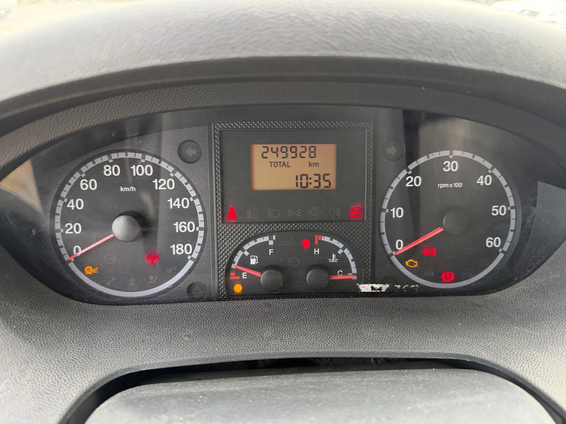 Zbliżenie na deski rozdzielcze samochodu. Po lewej stronie wskaźnik prędkości (0-180 km/h) z czerwoną igłą na 0 km/h, po prawej tachometr (0-60, oznaczony jako rpm x100) z czerwoną igłą na 0. W centralnym wyświetlaczu cyfrowym widoczny przebieg 249928 km (TOTAL km) oraz czas 10:35. Poniżej wyświetlacza wskaźniki paliwa (poziom blisko E) i temperatury chłodniczki (wskazanie między C a H). Aktywne czerwone lampki ostrzegawcze: pas bezpieczeństwa, bateria, silnik, ciśnienie oleju oraz żółta lampka kontrolna. Tło deski rozdzielczej szare, widoczny fragment kierownicy na górze.