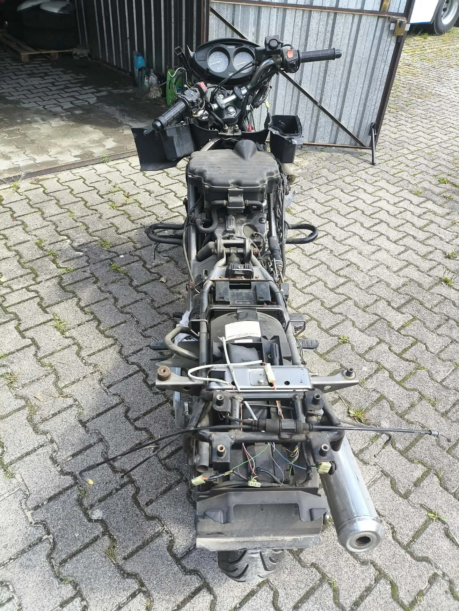 Zdjęcie przedstawia ramę motocykla bez nadwozia, ustawioną pionowo na kostce brukowej. Widoczny jest kierownica z zegarami, bak paliwa, silnik oraz tylna część ramy z wydechem. Motocykl jest w stanie rozbiórki, z widocznymi przewodami elektrycznymi i elementami konstrukcyjnymi.