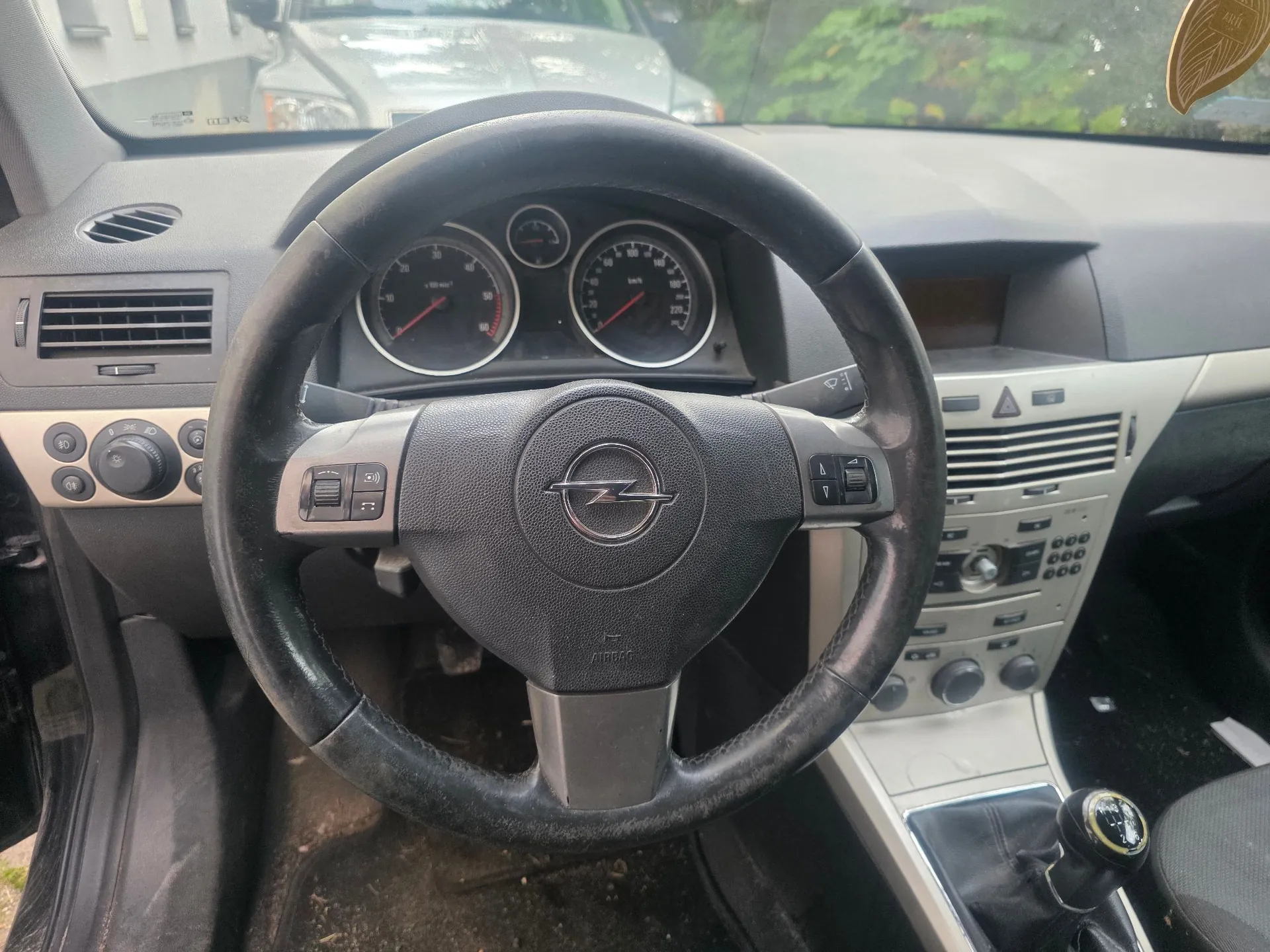 Wnętrze samochodu marki Opel, widok z perspektywy kierowcy. W centrum znajduje się czarna, skórzana kierownica z logo Opela na środku. Za kierownicą widoczna jest deska rozdzielcza z dwoma analogowymi wskaźnikami prędkości i obrotów oraz centralna konsola z radiem, przyciskami sterowania i lewarkiem zmiany biegów. Po lewej stronie widoczne są pokrętła klimatyzacji i nawiewy powietrza.