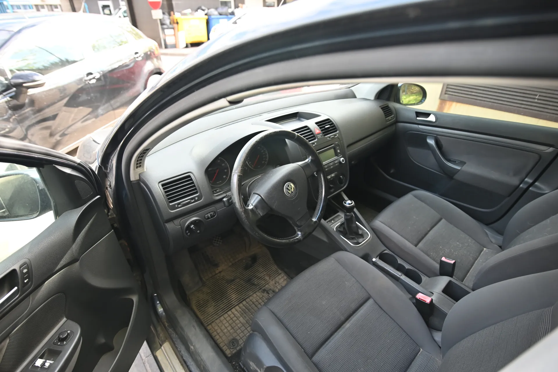 Wnętrze samochodu marki Volkswagen z otwartą kierowcą drzwiami. Widoczne czarne siedzenia, kierownica z logo VW, deska rozdzielcza z licznikami, dźwignia zmiany biegów oraz brudne dywaniki. Poza samochodem zaparkowane są inne pojazdy, żółty kontener i znak stop.