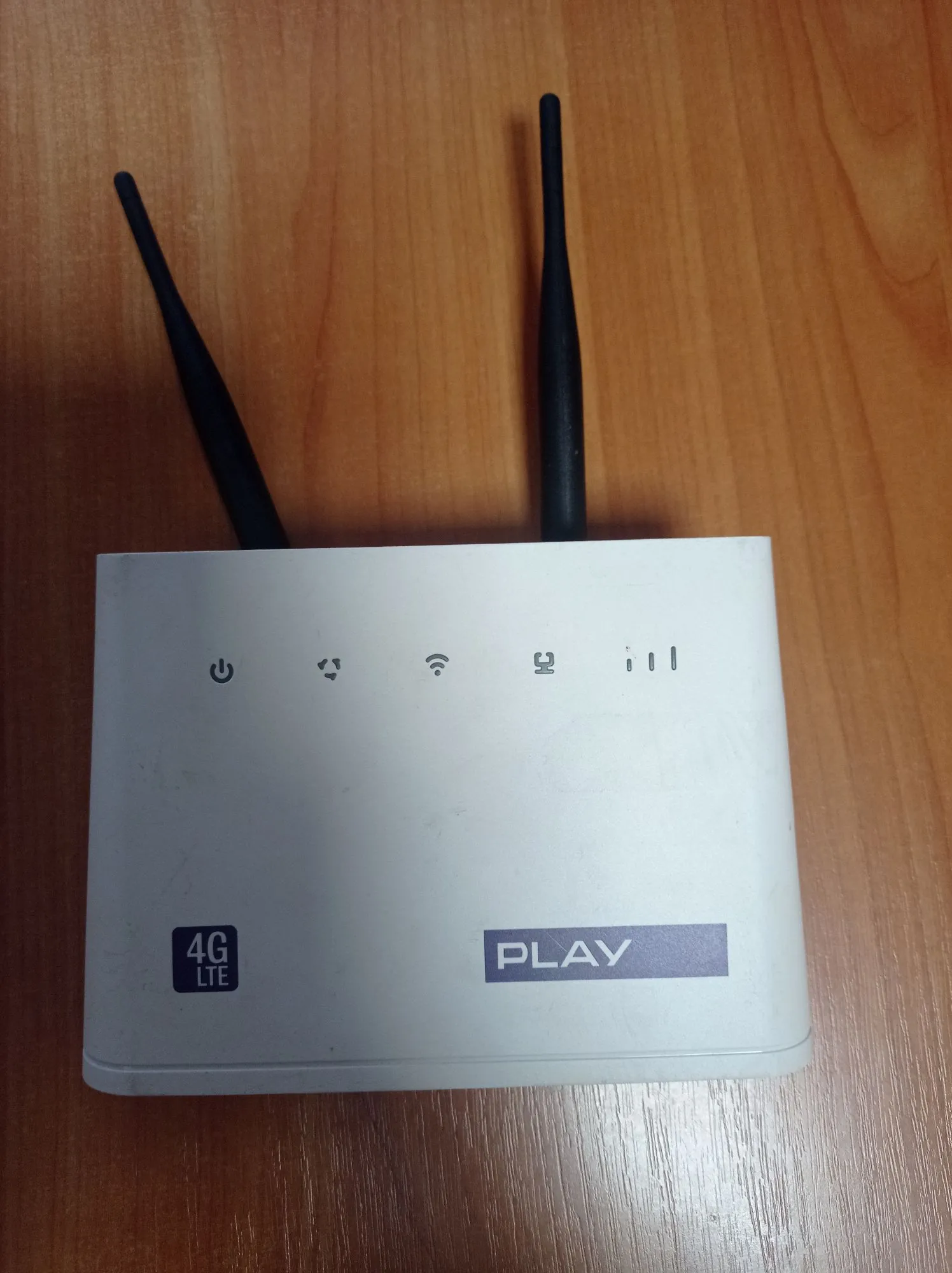 Biały router LTE marki Play z dwiema czarnymi antenami skierowanymi w górę, leżący na drewnianym blacie. Na przednim panelu urządzenia znajdują się ikony diod kontrolnych (zasilanie, sieć, Wi-Fi, sygnał) oraz logo 'PLAY' na niebieskim tle i oznaczenie '4G LTE'.