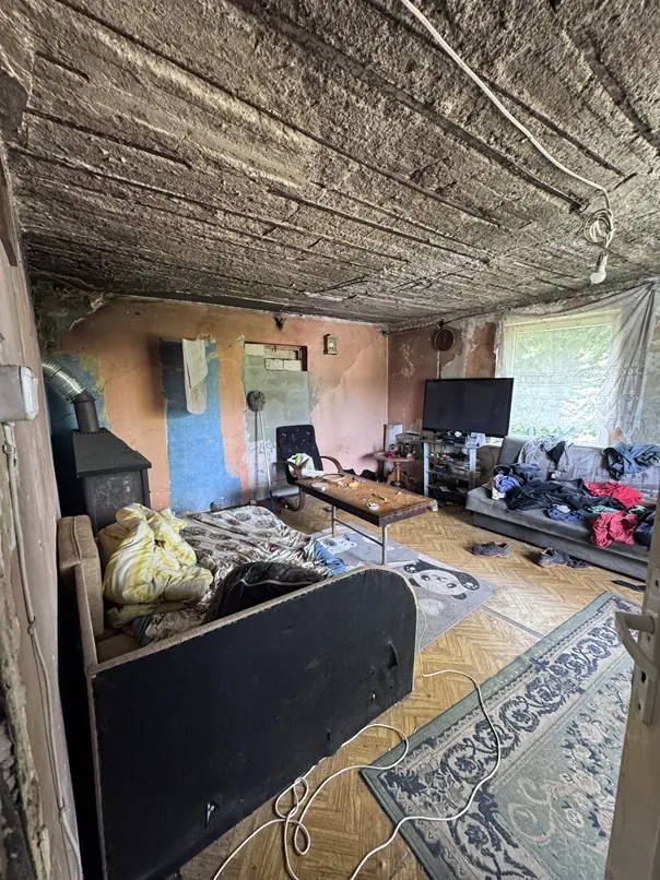Wnętrze zaniedbanego pomieszczenia z niskim, betonowym sufitem. W pokoju znajduje się łóżko z pościelą i ciemnym zagłówkiem, na którym leżą ubrania. Obok stoi mały, czarny piecyk. W głębi pomieszczenia widoczny jest drewniany stolik kawowy, telewizor na szafce oraz szara kanapa zasypana ubraniami. Ściany są w złym stanie, z odpadającym tynkiem i widocznymi plamami farby w kolorach różowym i niebieskim. Na podłodze z parkietu leżą dwa dywany, a przez okno wpada światło dzienne.
