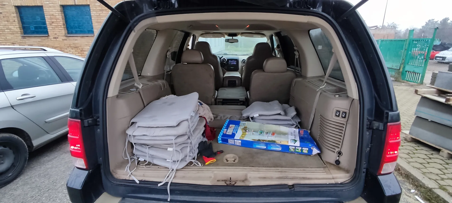 Otwarty bagażnik czarnego SUV-a z beżowym wnętrzem. W bagażniku znajdują się stosy szarych poduszek lub mat z białymi sznurkami, niebieskie pudełko z grafiką i tekstem, czarne i czerwone rękawice oraz czerwony przedmiot. W tle widoczny srebrny samochód, ceglany budynek z niebieskimi oknami, zielone ogrodzenie i stosy materiałów na paletach. Podłoga wyłożona cegłą.