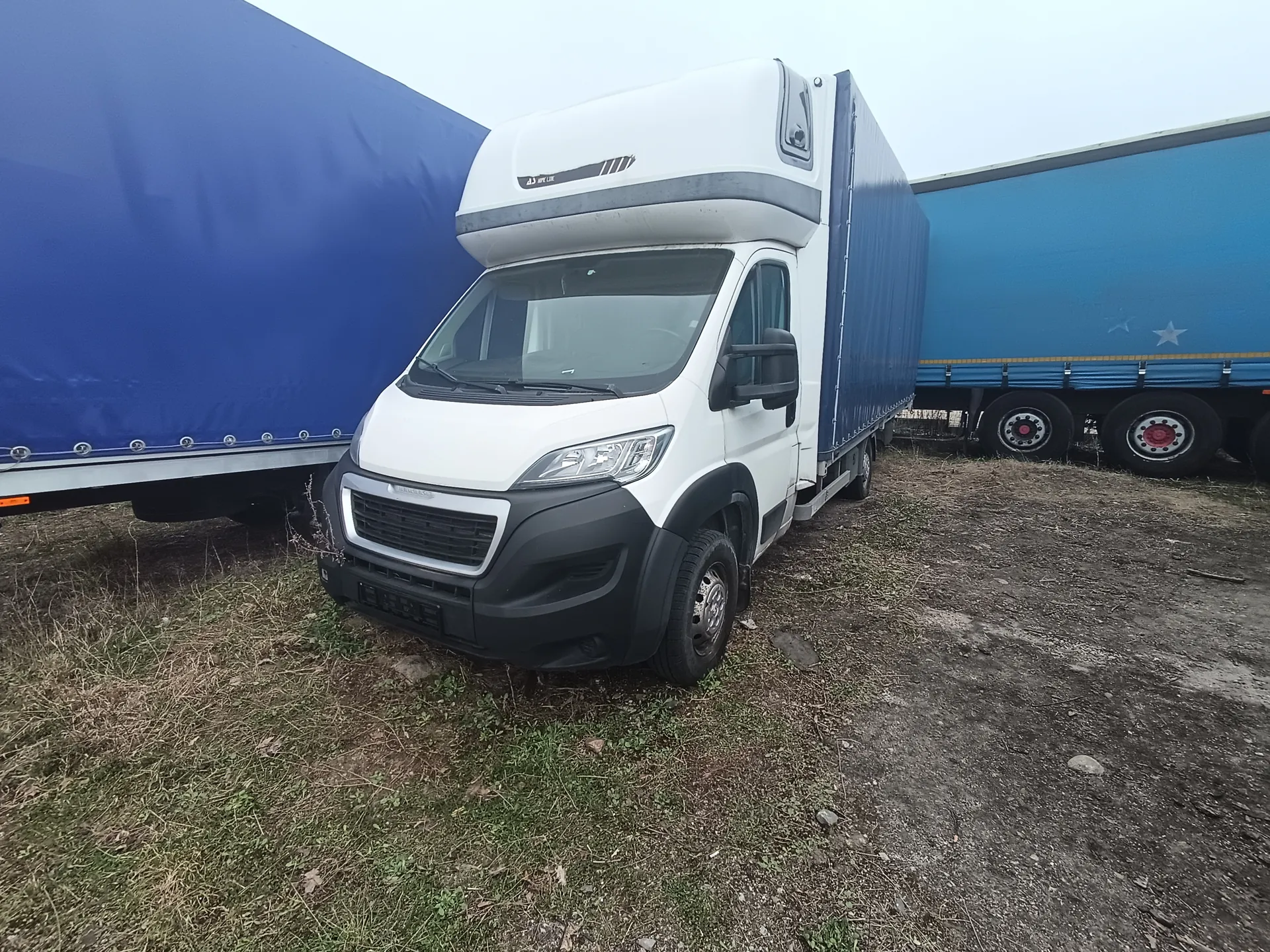 Biały samochód dostawczy z niebieskim nadwoziem przyczepy zaparkowany na trawiastym i ziemnym terenie. W tle dwa duże niebieskie przyczepy ciężarowe. Niebo jest pochmurne. Samochód posiada czarną maskę przednią, reflektory oraz widoczne koła. Na przyczepach widoczne są elementy metalowe i koła z czerwonymi śrubami.