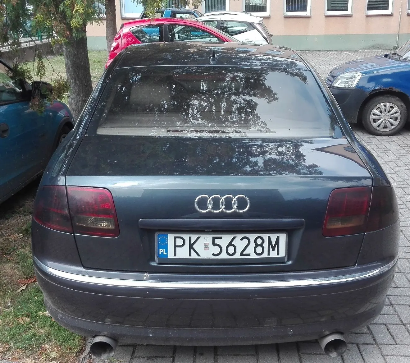 Tylny widok ciemnoszarego samochodu marki Audi z tablicą rejestracyjną PK 5628M. Samochód zaparkowany jest na kostce brukowej, w tle widoczne są inne zaparkowane pojazdy: czerwony i niebieski, oraz fragment budynku i drzewo.