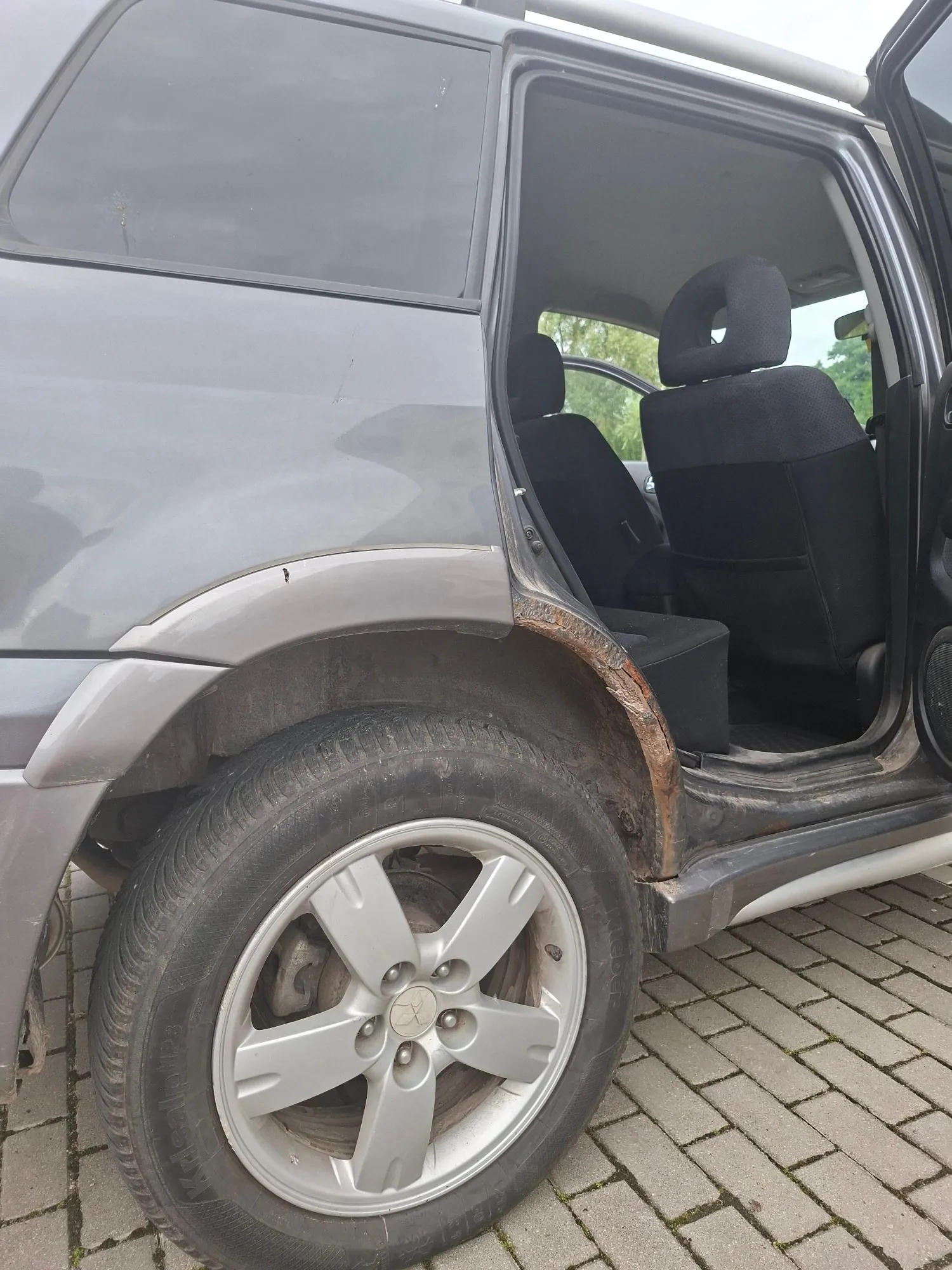 Zdjęcie fragmentu srebrnego samochodu, pokazujące przednie prawe koło z pięcioramienną aluminiową felgą oraz widoczną korozję na nadkolu nad oponą. W tle widać otwarte drzwi kierowcy i czarne siedzenie samochodu oraz kostkę brukową na ziemi.