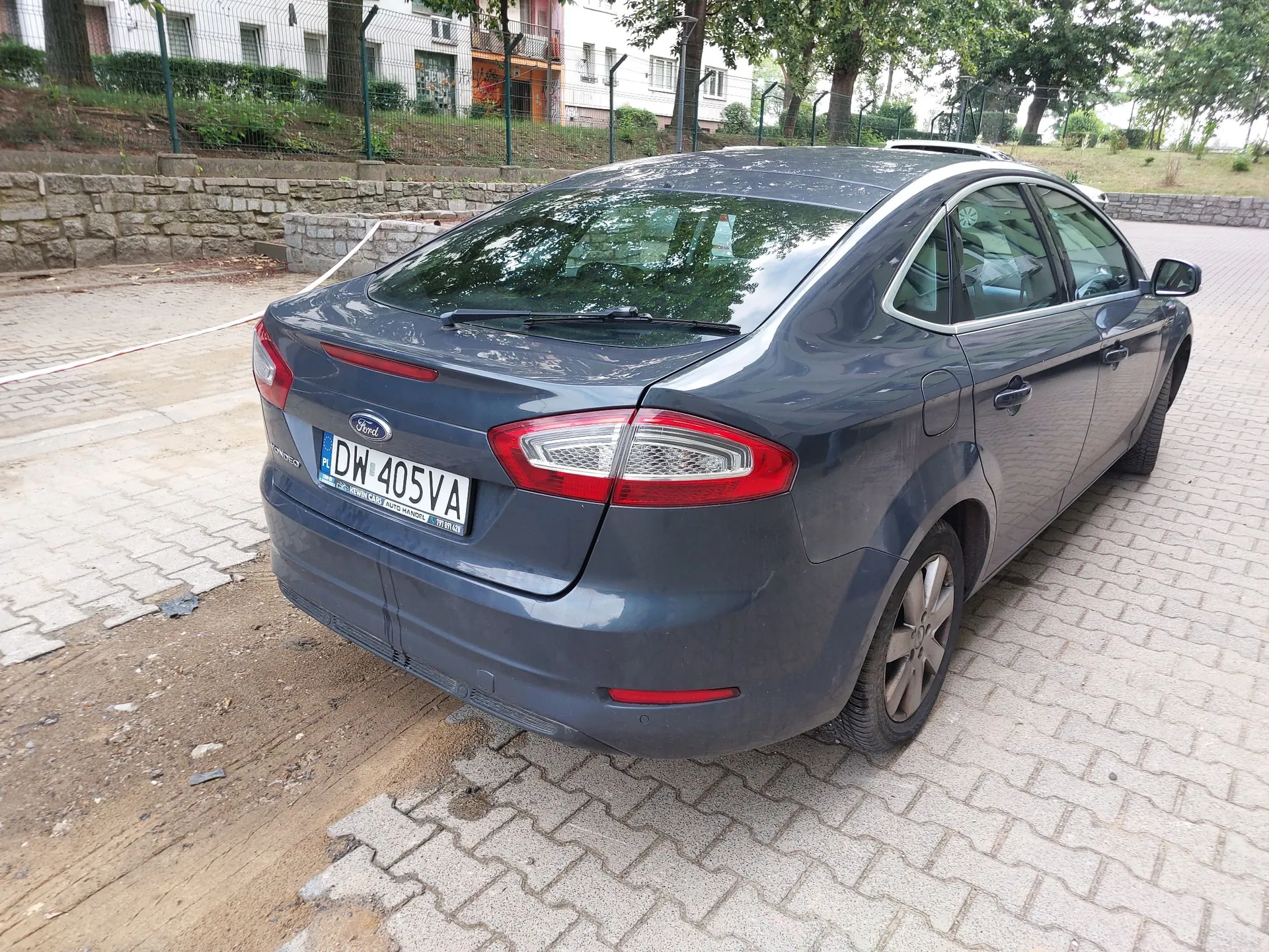 Tylny widok ciemnoszarego samochodu marki Ford zaparkowanego na kostce brukowej. Pojazd posiada polską tablicę rejestracyjną o numerze DW 405VA. W tle widoczny jest niski kamienny murek, zieleń oraz fragmenty budynków mieszkalnych.