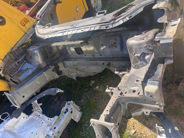Zdjęcie przedstawia rozłożone metalowe elementy karoserii samochodowej, w tym fragmenty ramy i paneli, leżące na trawie i ziemi. W tle widoczny jest żółty pojazd, prawdopodobnie maszyna budowlana, z częściowo widocznym kołem. Elementy karoserii mają uszkodzenia, otwory i ślady korozji. Brak czytelnego tekstu na obrazie.