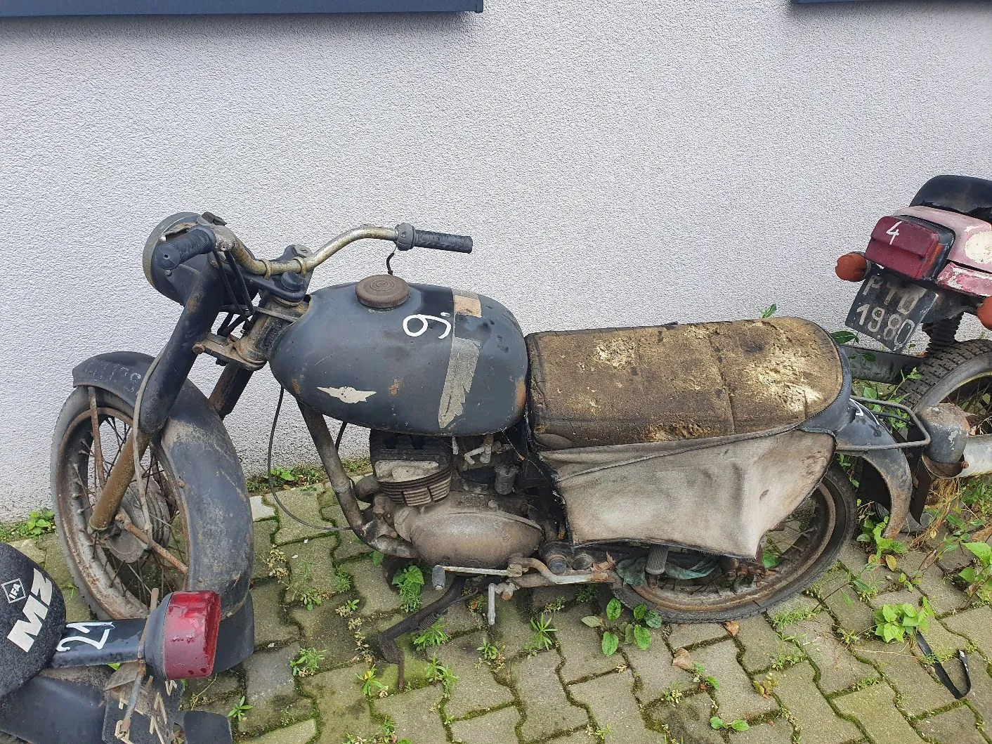 Stary, zardzewiały motocykl w kolorze czarnym, zaparkowany na kostce brukowej z wyrastającą między szczelinami trawą. Motocykl jest w złym stanie technicznym, z widocznymi śladami korozji i zabrudzeniami na baku oraz siedzeniu. Na baku widnieje biała cyfra '9'. W tle znajduje się jasna, chropowata ściana budynku. Po prawej stronie widoczny jest fragment innego pojazdu z tablicą rejestracyjną, na której widnieją znaki '19.80'.