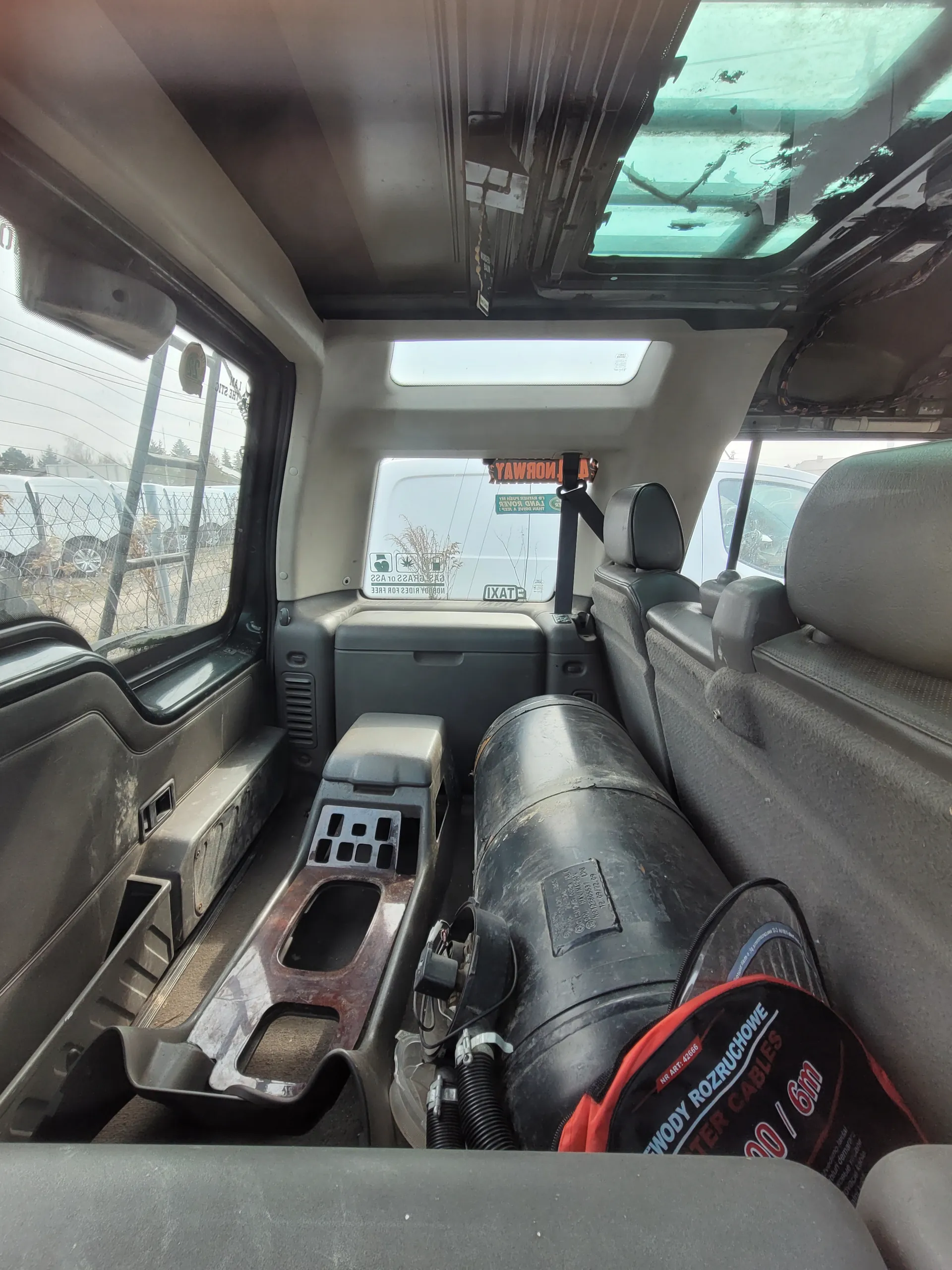Wnętrze pojazdu, prawdopodobnie furgonetki lub ciężarówki. Widoczne są siedzenia po prawej stronie, duże czarne cylindryczne zbiorniki z napisami "ROZRUCHOWE", "10/6m" oraz "CABLES". Na jednym z zbiorników znajduje się czerwono-czarna etykieta z białym tekstem. W tle przez okna widać zewnętrzne elementy: siatkę ogrodzeniową, inne pojazdy i drzewa. Na przedniej szybie umieszczono pomarańczowy znak z napisem "PROBAYA" oraz zieloną naklejkę z symbolami. Wnętrze posiada beżowe i szare elementy wykończeniowe.