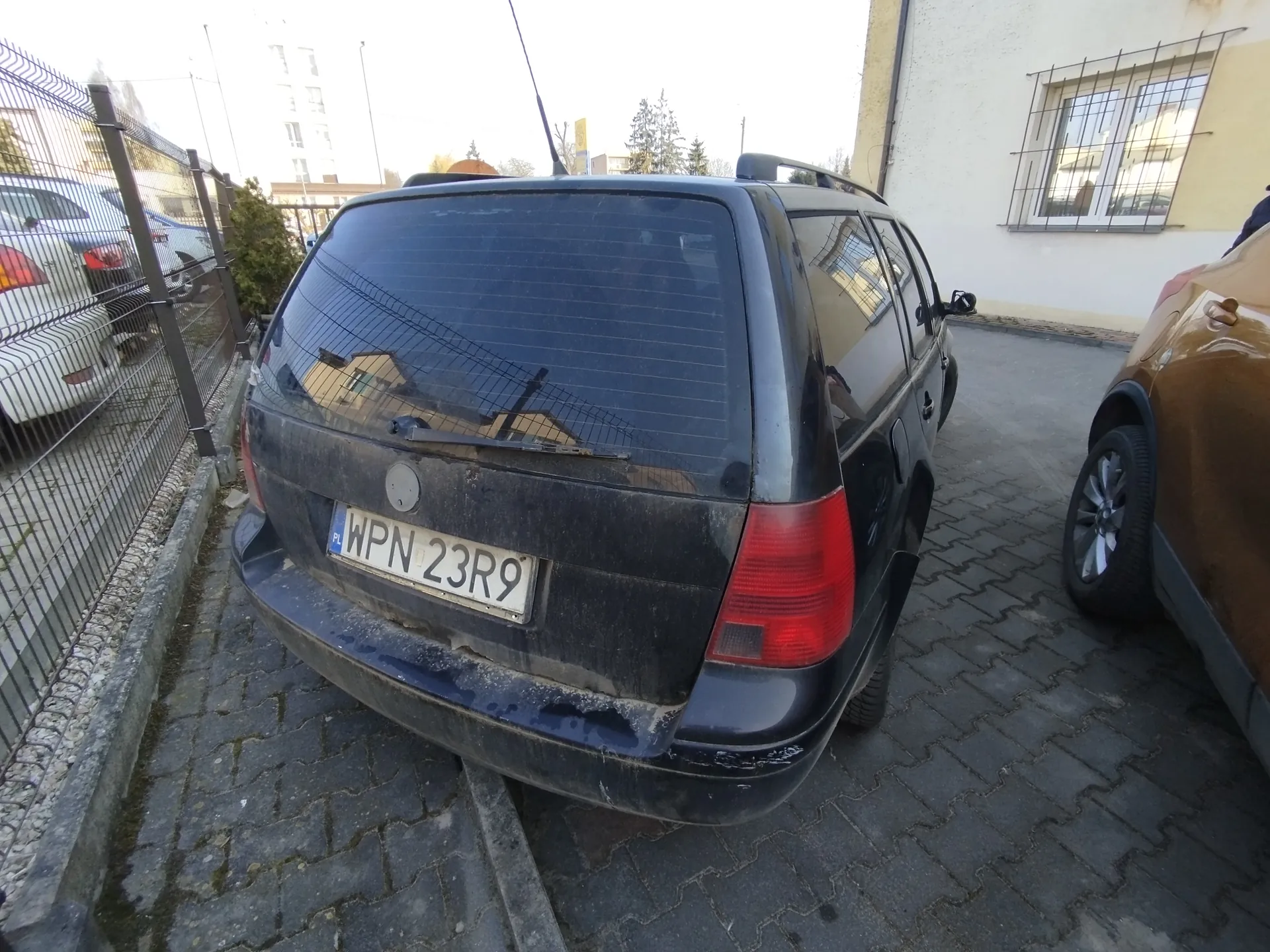 Czarny samochód typu kombi zaparkowany na brukowanej powierzchni, widok od tyłu. Na tylnej szybie odbicie siatki ogrodzenia i budynków. Tablica rejestracyjna z czytelnym napisem PL WPN 23R9. Samochód pokryty brudem, widoczne zadrapania na zderzaku. Po lewej stronie metalowe ogrodzenie z innym białym samochodem za nim, po prawej stronie częściowo widoczny brązowy samochód. W tle budynki mieszkalne, drzewa i jasne niebo.